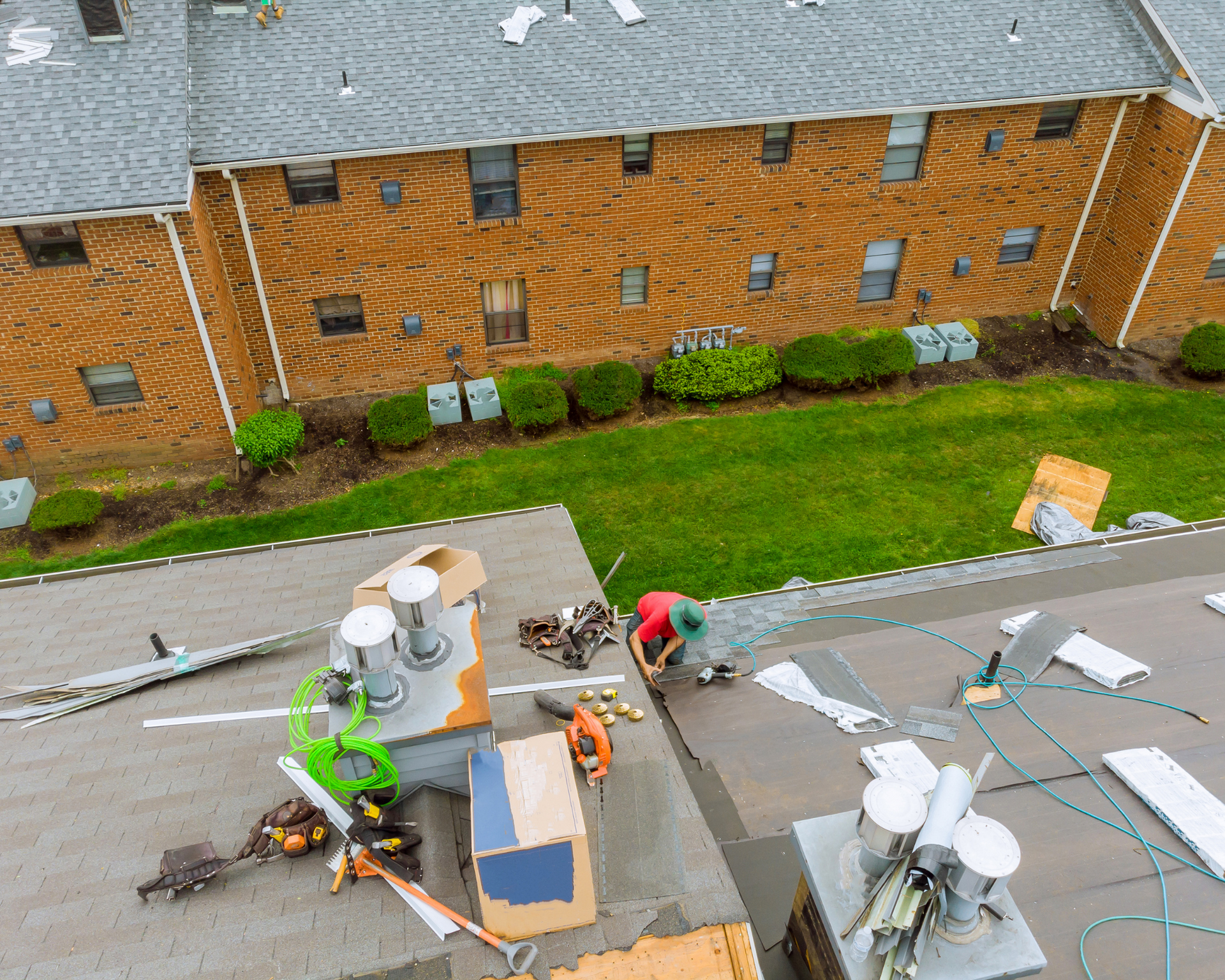 Roofer working on a flat roof with tools and materials. Apartments with brick siding and green lawn in the background.