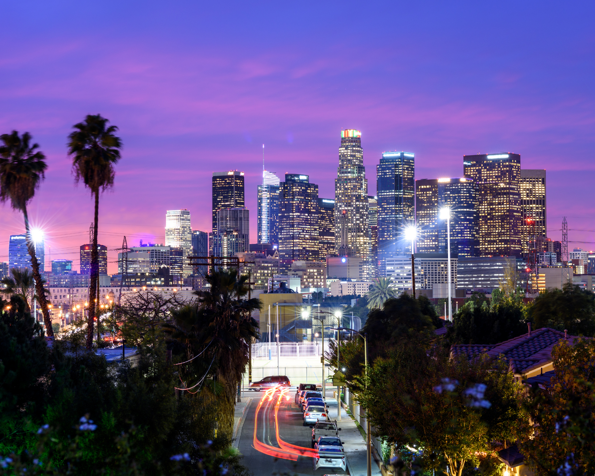 Los Angeles skyline at dusk, buildings aglow against a purple sky. Palm trees and a street with car lights in the foreground.