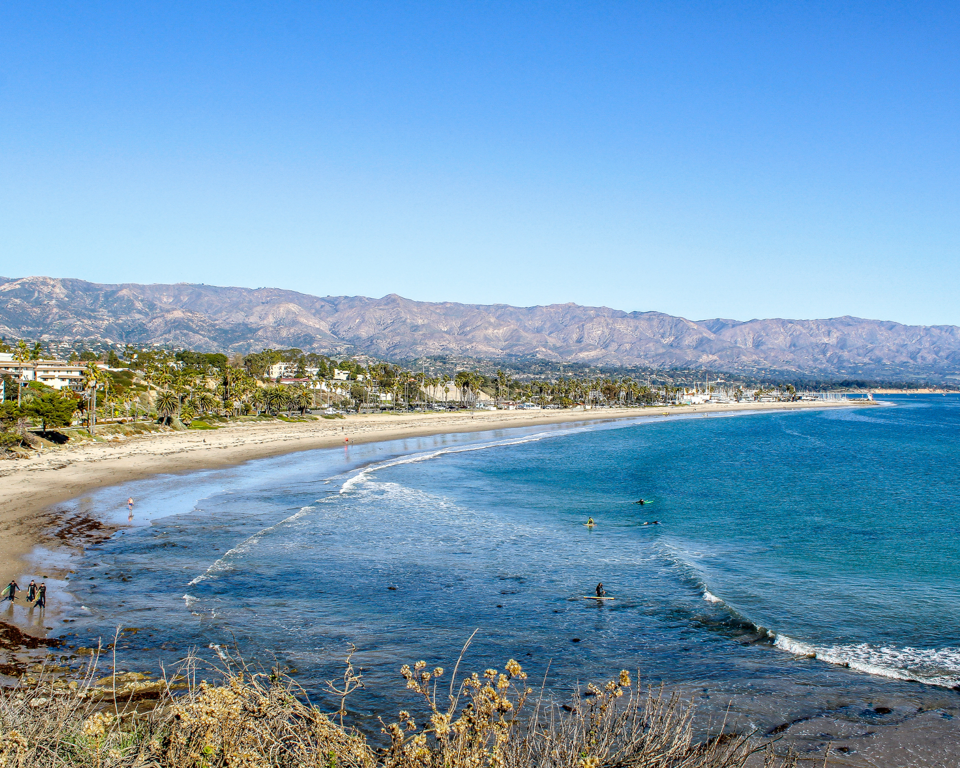 Sandy beach with turquoise water, low waves, and a mountain range under a clear blue sky.