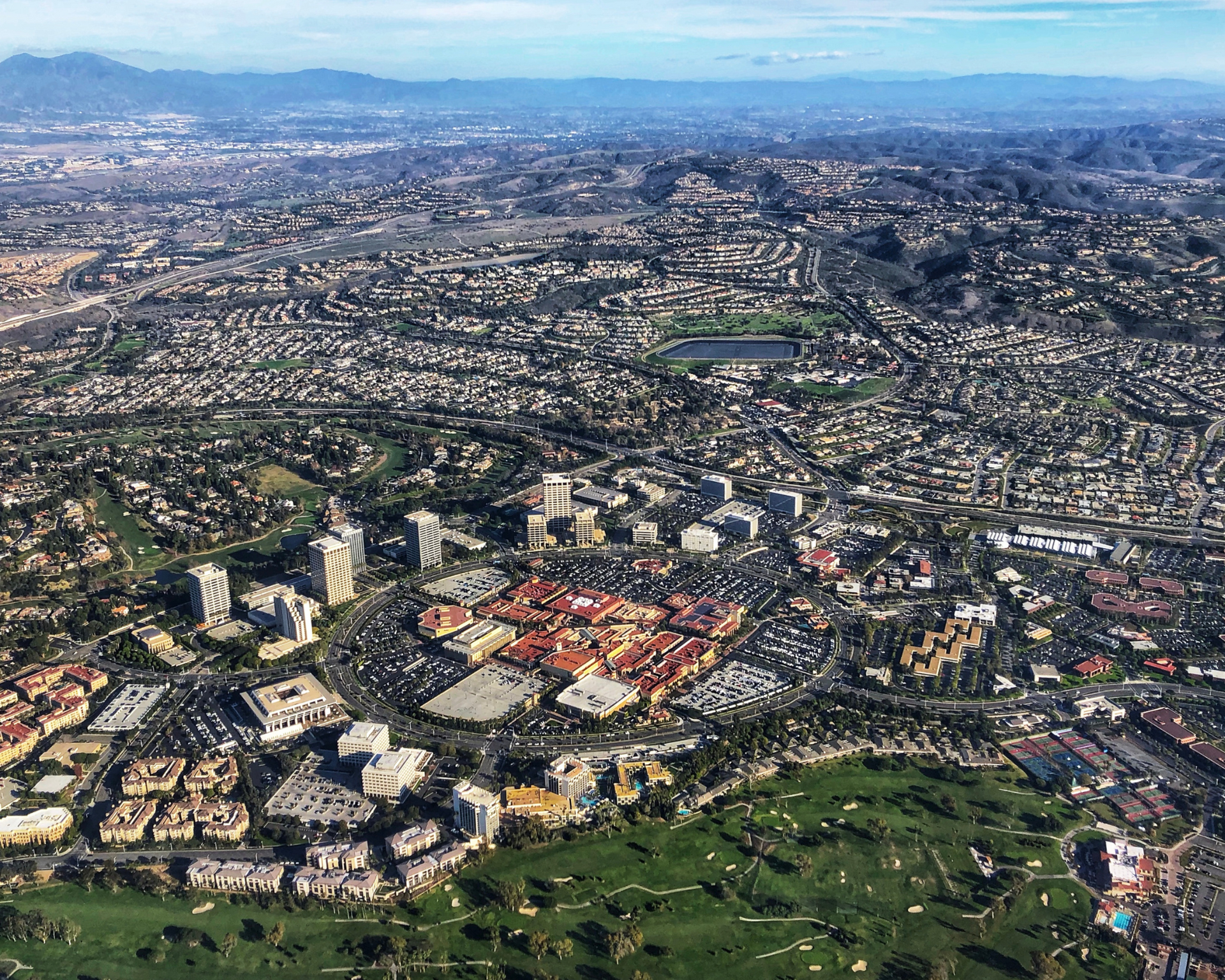 Aerial view of a sprawling urban area with buildings, roads, a shopping center, and a golf course under a blue sky.