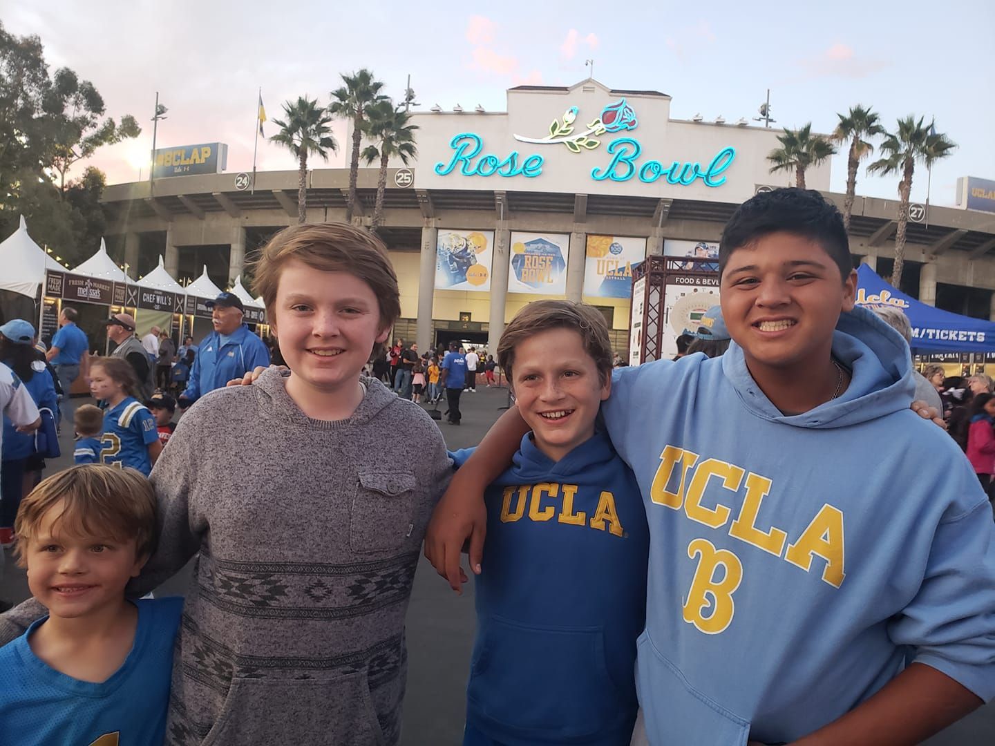 Four people in front of the Rose Bowl. Three in UCLA apparel smile; one boy has his arm around another.