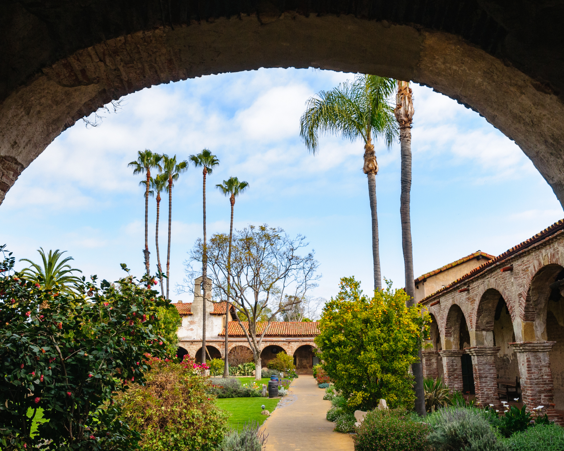 Courtyard view through an arched opening: palm trees, walkway, arched colonnade, greenery, blue sky.