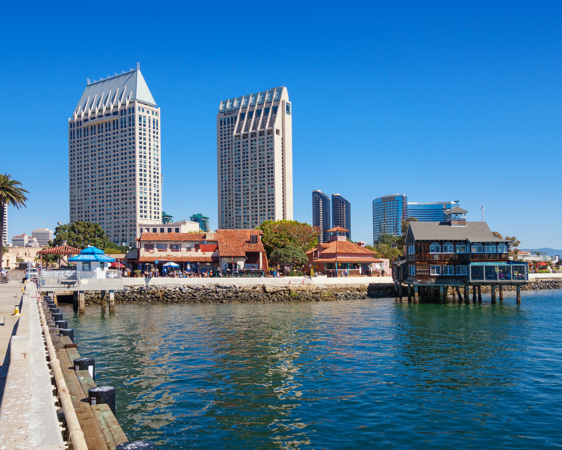 Waterfront buildings in San Diego with two tall towers, shops, blue water, and clear sky.
