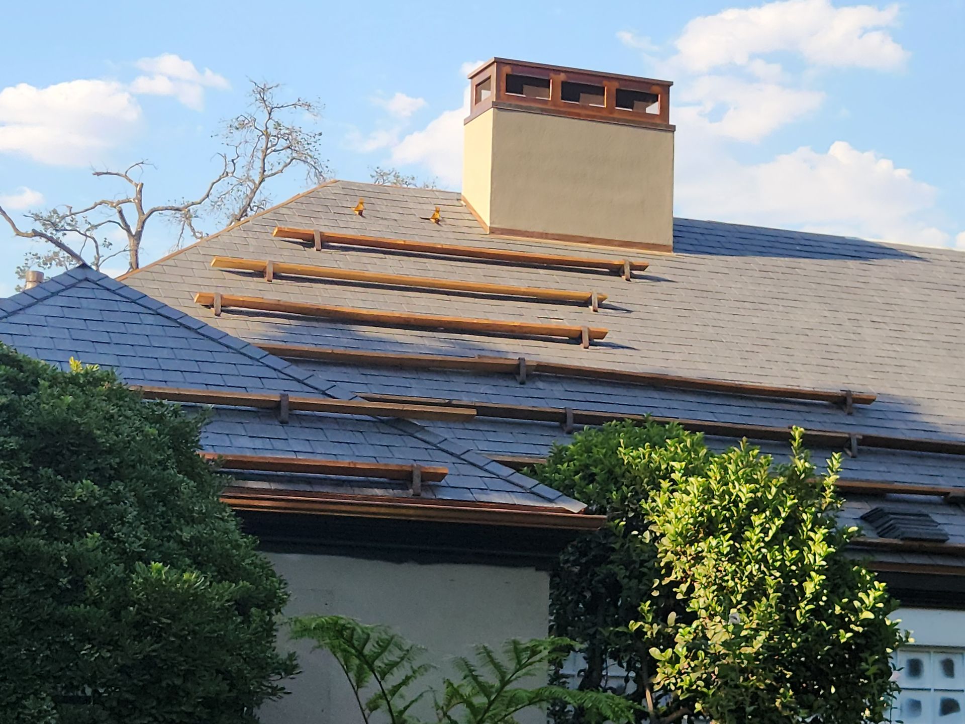 A house roof with scaffolding, chimney, and gray shingles. Green bushes and a blue sky are visible.