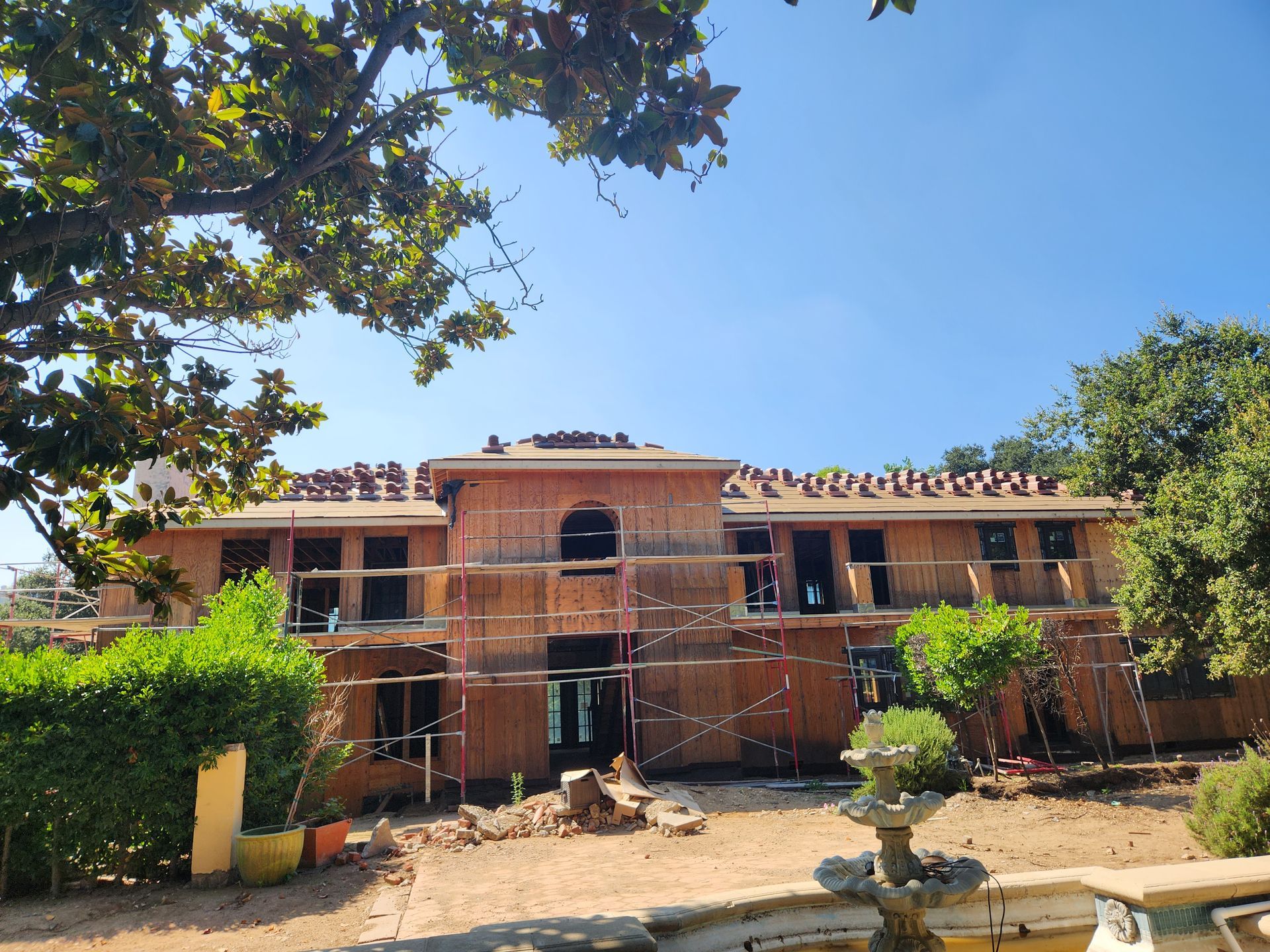 House under construction, brown scaffolding, missing windows, and roof tiles. Exterior view under a blue sky.