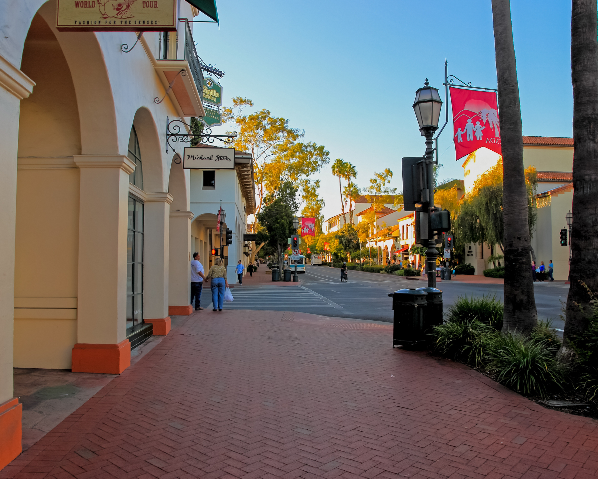 Brick-paved street lined with shops, arches, and palm trees. People walking. Flags and streetlights in sunny California setting.