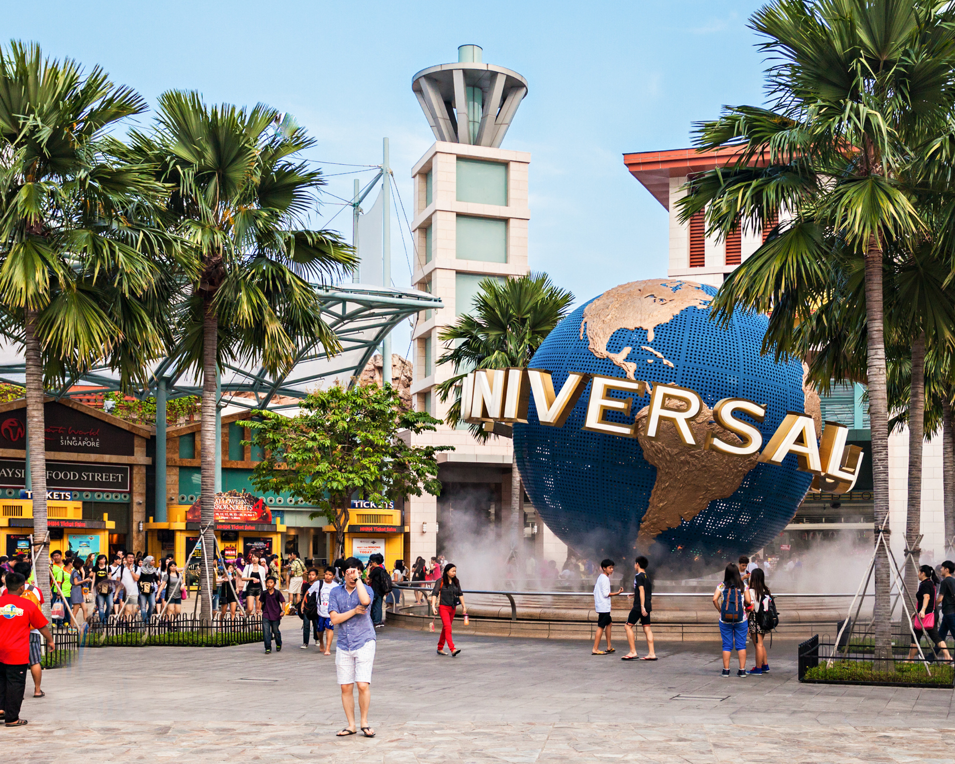 Entrance to Universal Studios Singapore, iconic globe, fountain, people, palm trees, sunny day.