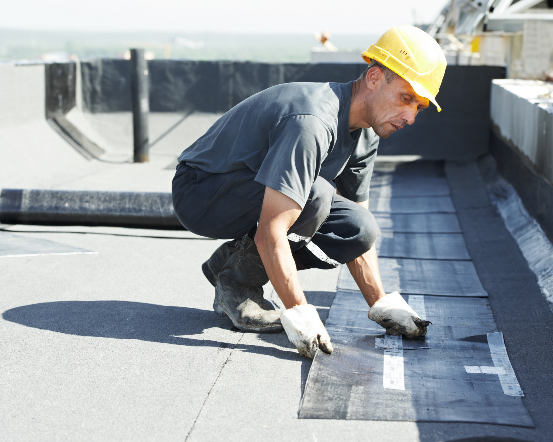 Roofer in yellow hardhat, installing dark roofing material on a flat roof.