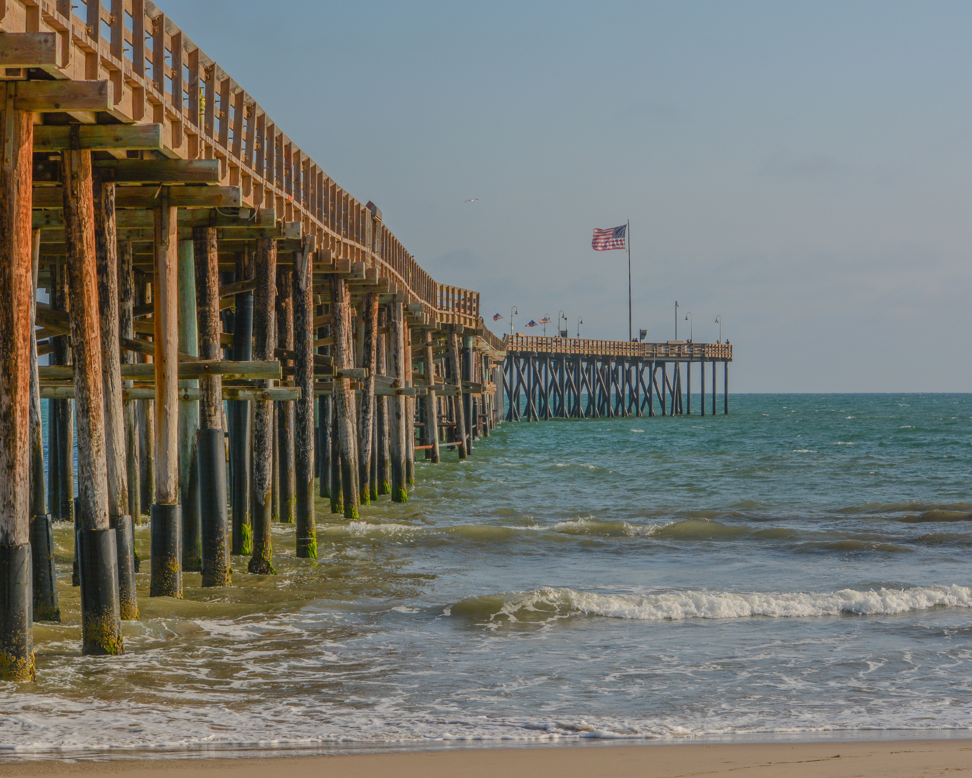Wooden pier extending into ocean; American flag waves atop the pier; waves crash on the sandy shore.