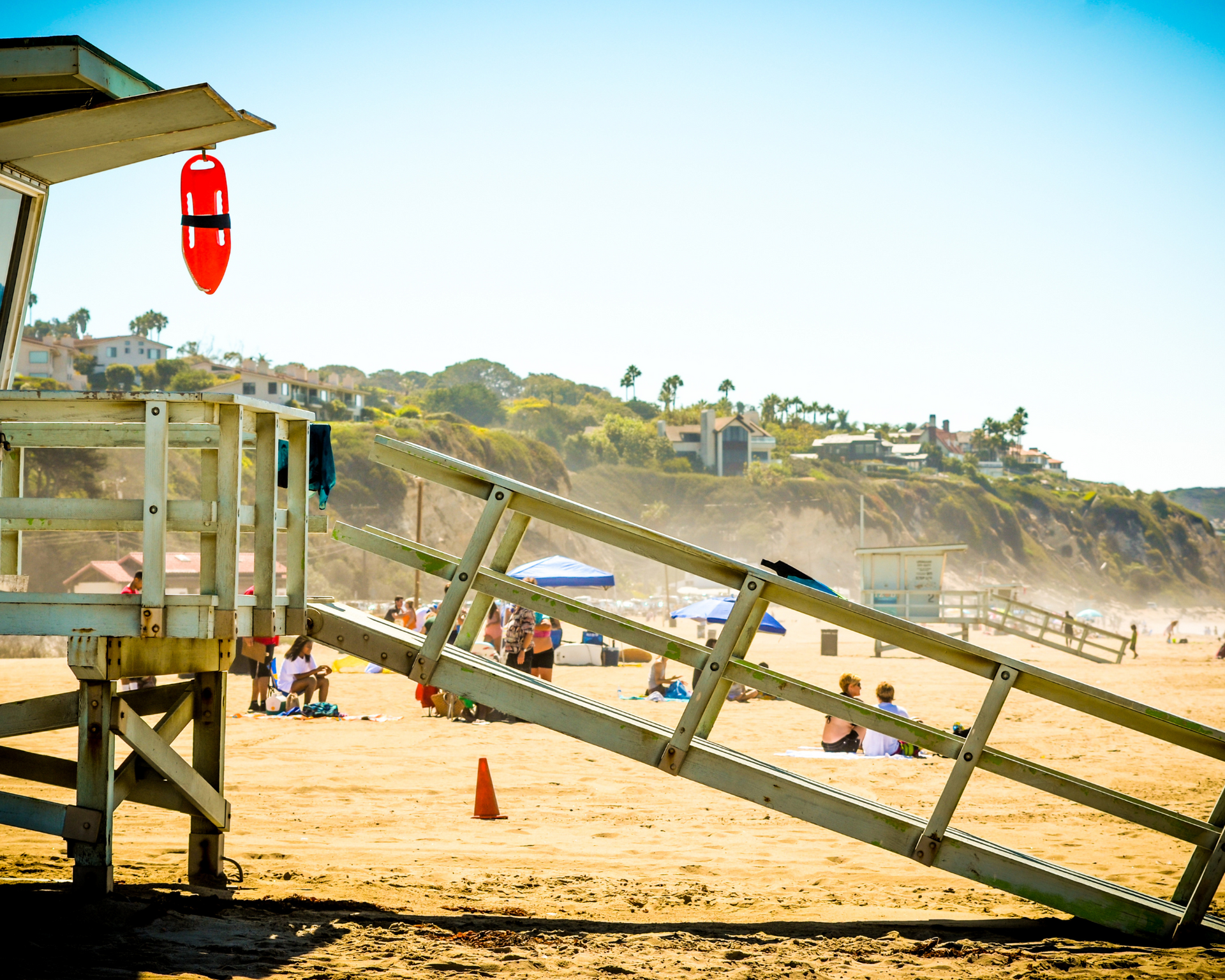 Lifeguard stand on a sunny beach, with a rescue buoy hanging. People relax in the sand and hillside homes in the distance.