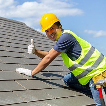 Roofer in safety gear on a tiled roof gives a thumbs-up against a blue sky.