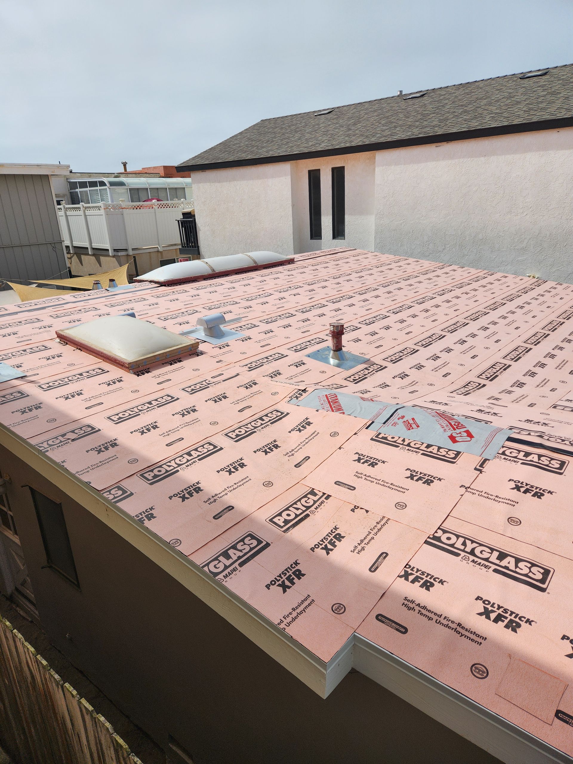 Pink insulation on a flat roof with a skylight and vent pipes, under construction with a white building in the background.