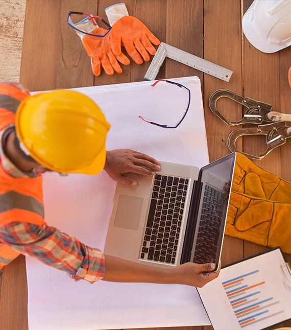 Construction worker at a desk with a laptop, blueprints, and safety equipment.