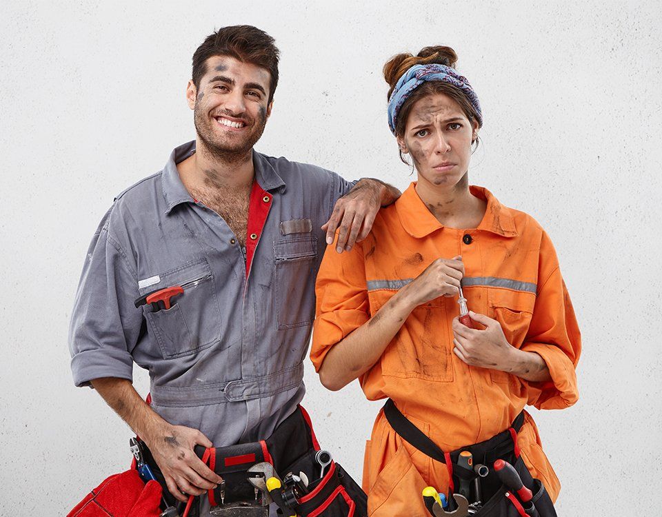 Man and woman in work coveralls, both covered in dirt. Man smiles; woman looks disgruntled. White background.