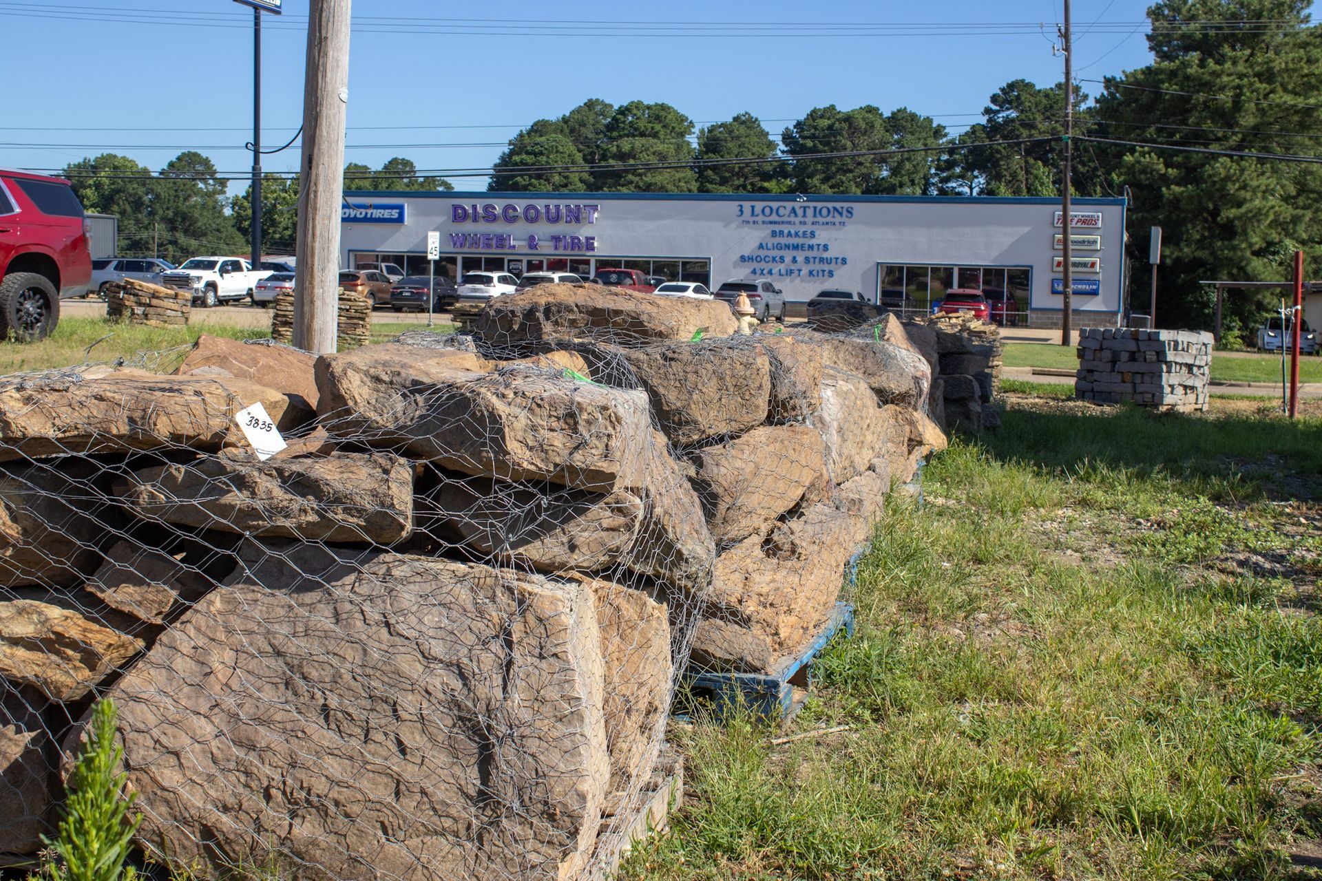 Large stacked stone sign in a grassy roadside lot with buildings and parked cars in the background