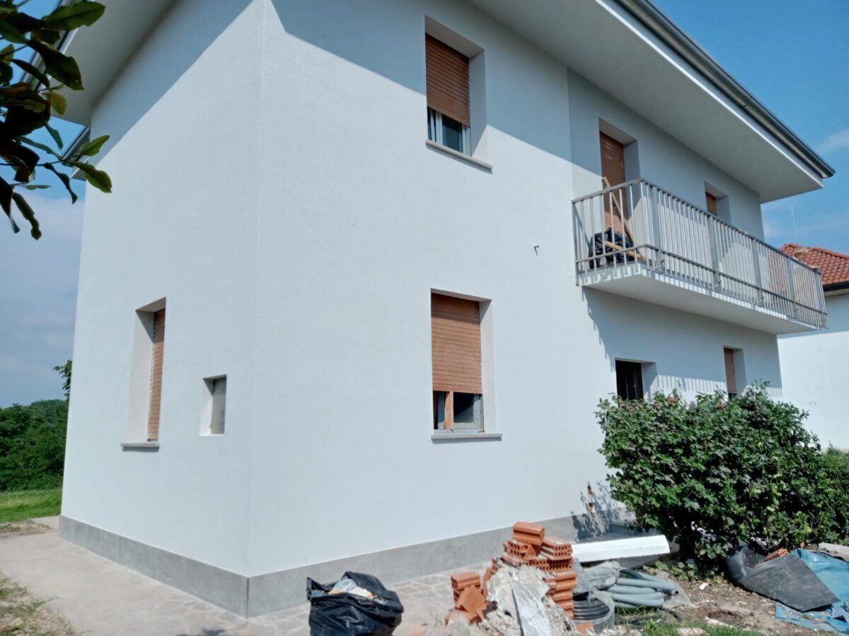 Casa bianca a due piani con persiane marroni chiuse e un piccolo balcone. Detriti di costruzione davanti.