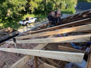 Worker on a steep roof framing, with lumber and a white truck below amid trees.