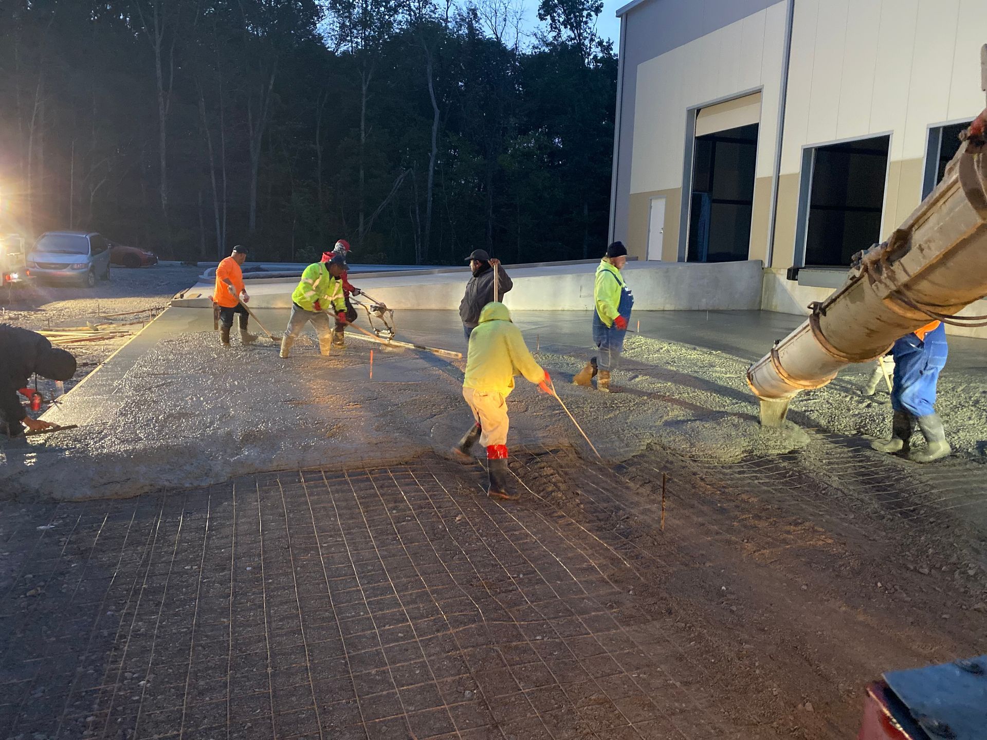 Road crew spraying water and spreading gravel on a wet pavement outside a warehouse at dusk