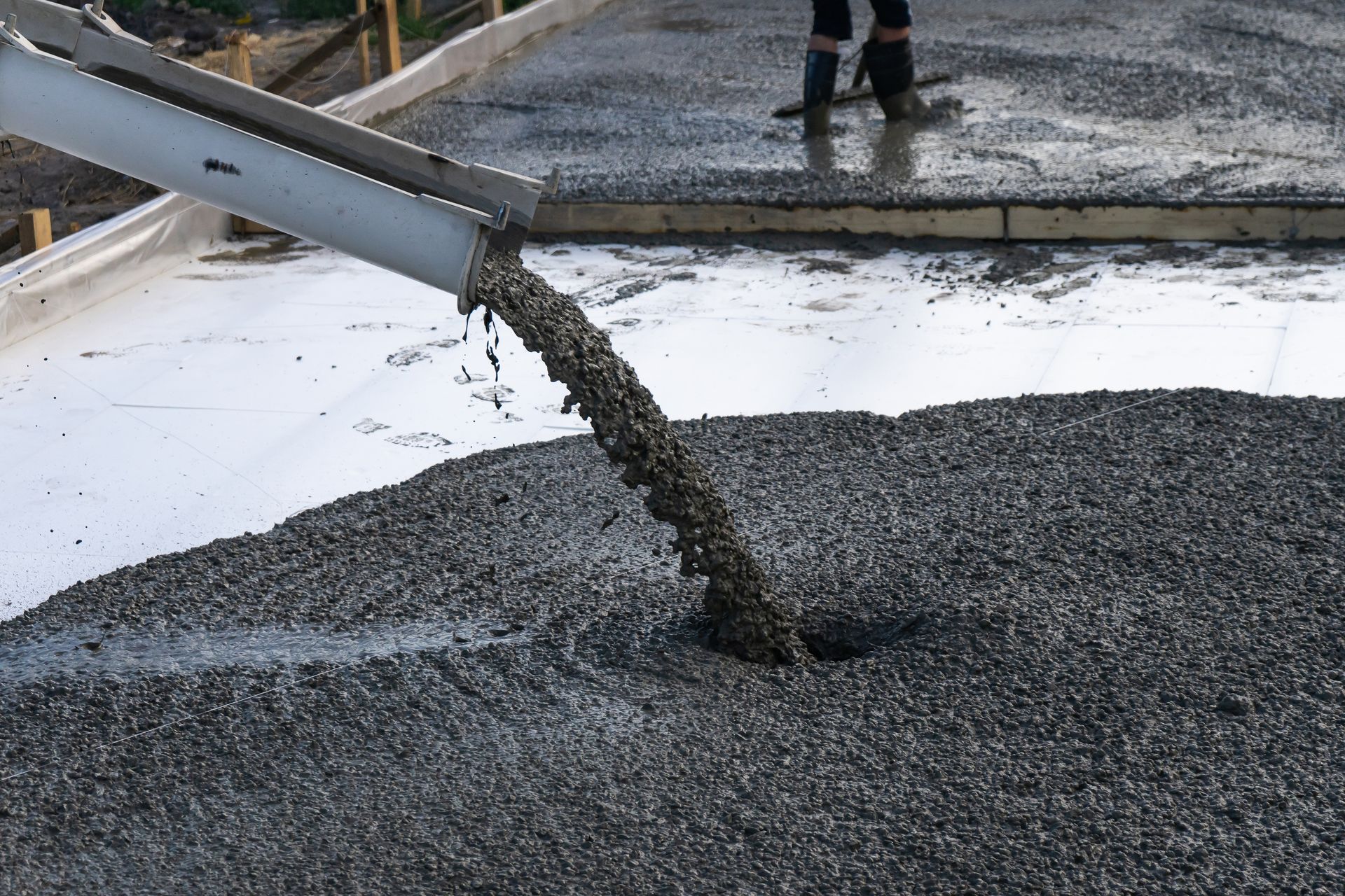 Wet concrete pours from a chute onto a construction site with a worker standing nearby.
