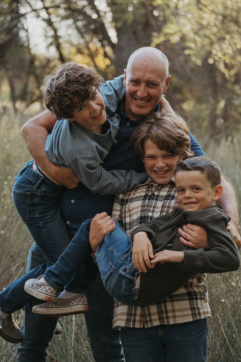 Three people carrying a smiling older man outdoors in a wooded area