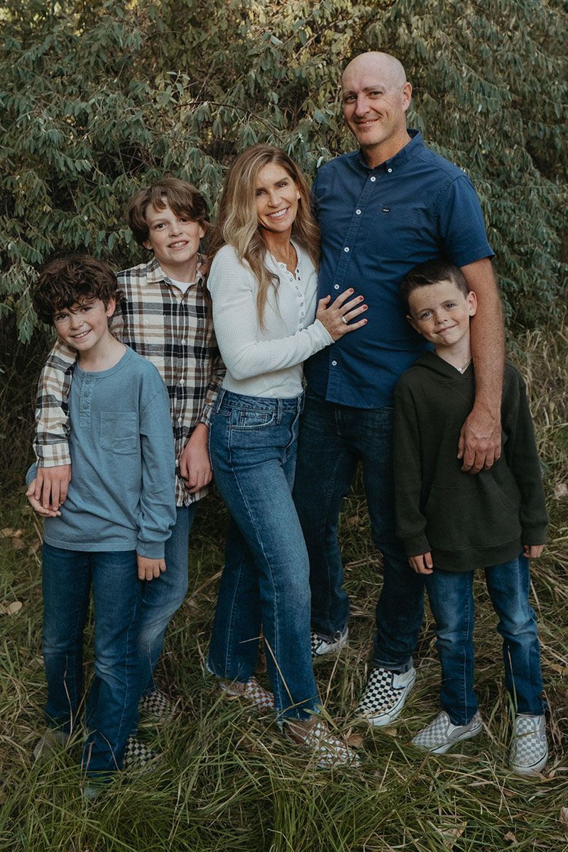 A family of four stands together outdoors in tall grass, smiling at the camera.