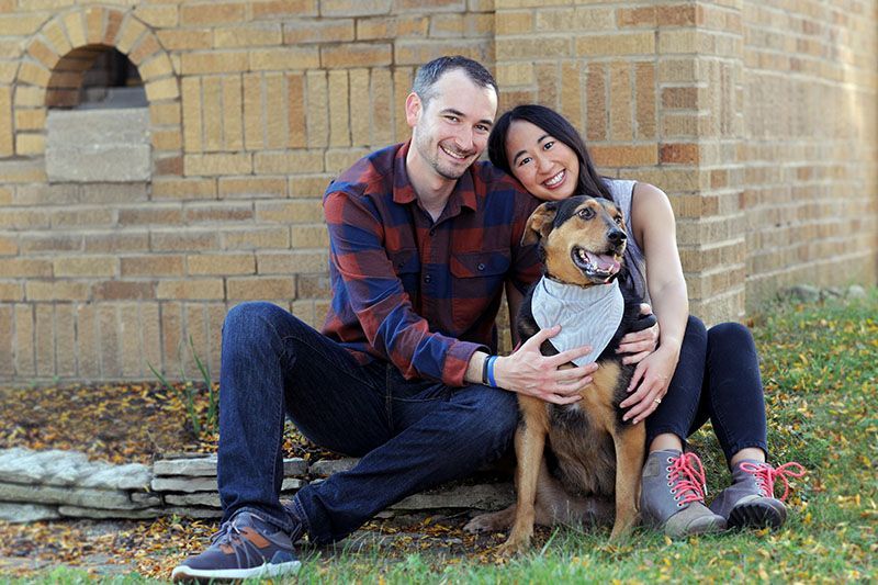 Couple sitting on grass with a dog in front of a brick wall, smiling at the camera