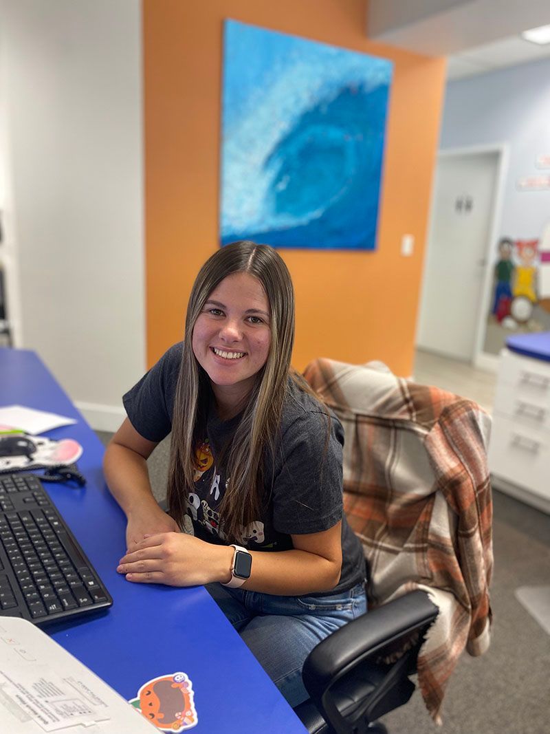 Smiling woman seated at a desk in an office, with a keyboard and orange wall art behind her