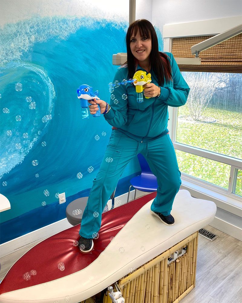 A person in teal scrubs stands on a surfboard-shaped table, holding bubble-blowing toy guns against a blue ocean mural.