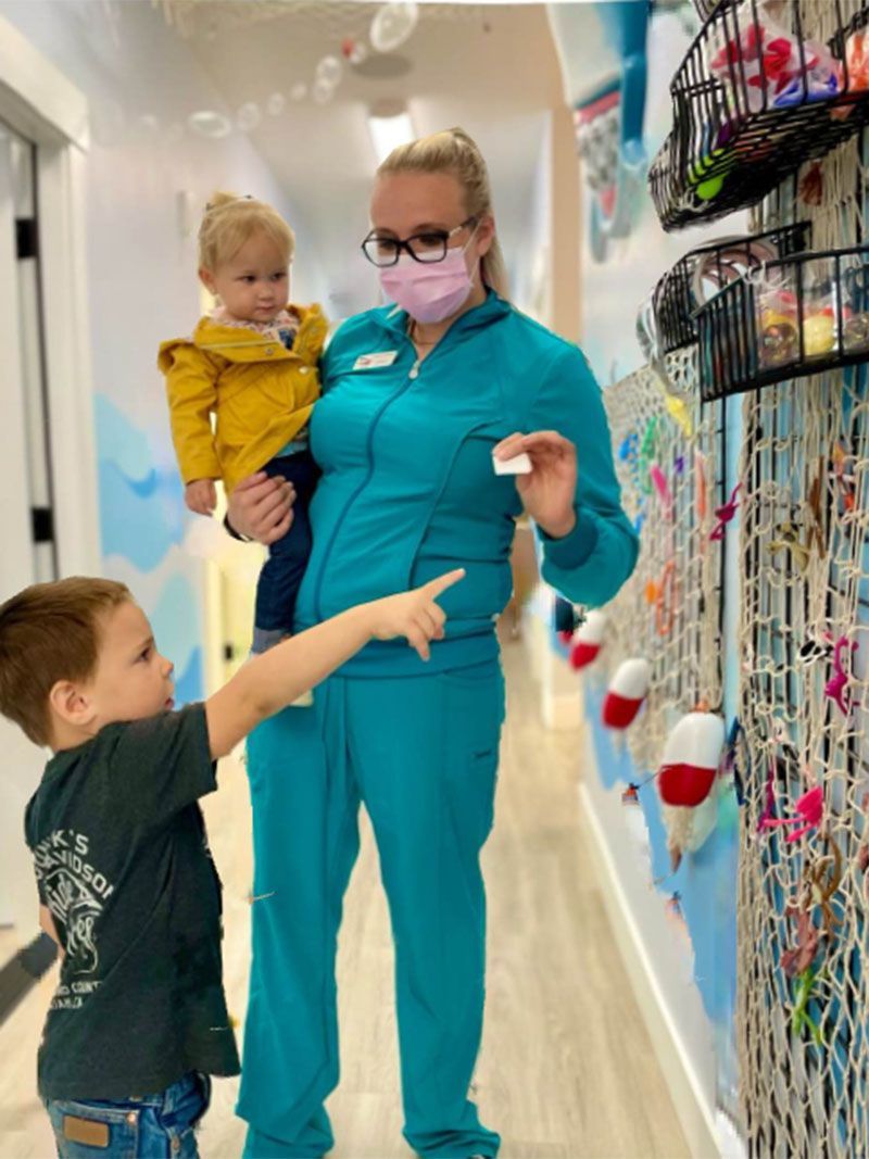Nurse in teal scrubs holding a toddler while a young child points at colorful wall displays in a hallway
