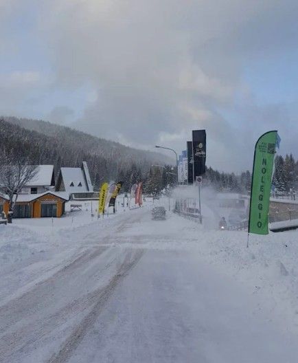 Strada innevata di una stazione sciistica con bandiere, baite e colline sotto un cielo nuvoloso.