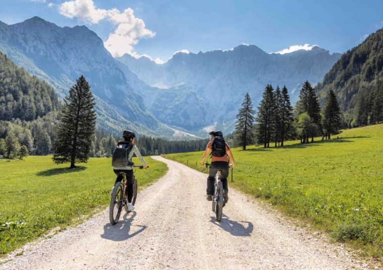 Due ciclisti percorrono un sentiero sterrato attraverso una verde vallata di montagna sotto un cielo azzurro brillante.