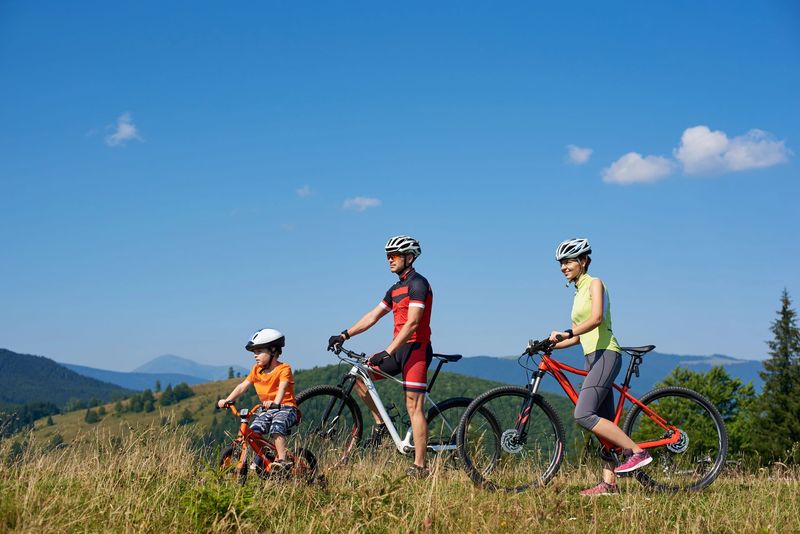 Tre ciclisti con il casco si fermano in sella alle loro mountain bike in un prato sotto un cielo azzurro.