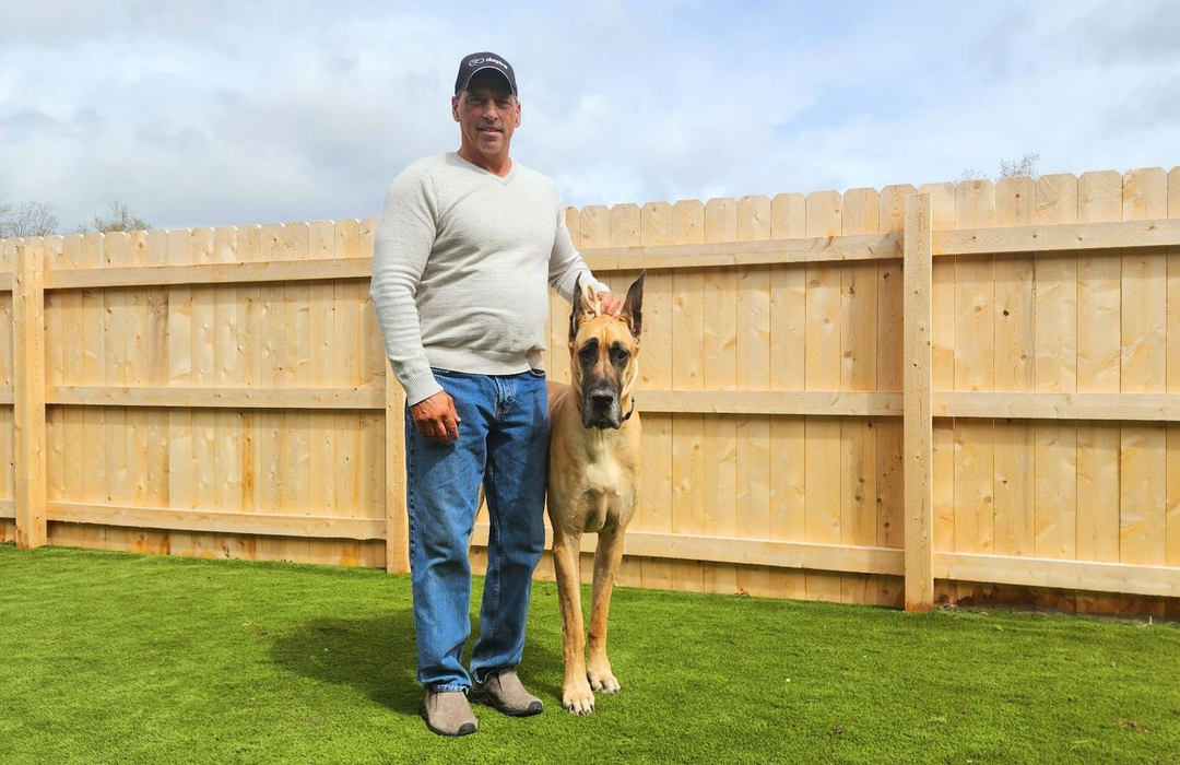 Man standing beside a tan dog in a grassy backyard with a wooden fence and cloudy sky