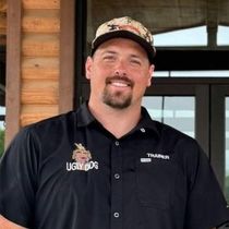 Man in black shirt and camo cap smiling outdoors in front of a wooden building