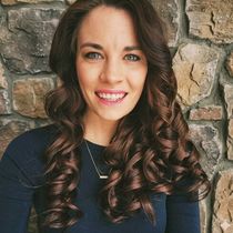 Smiling woman with curled brown hair in a navy top, standing before a stone wall.