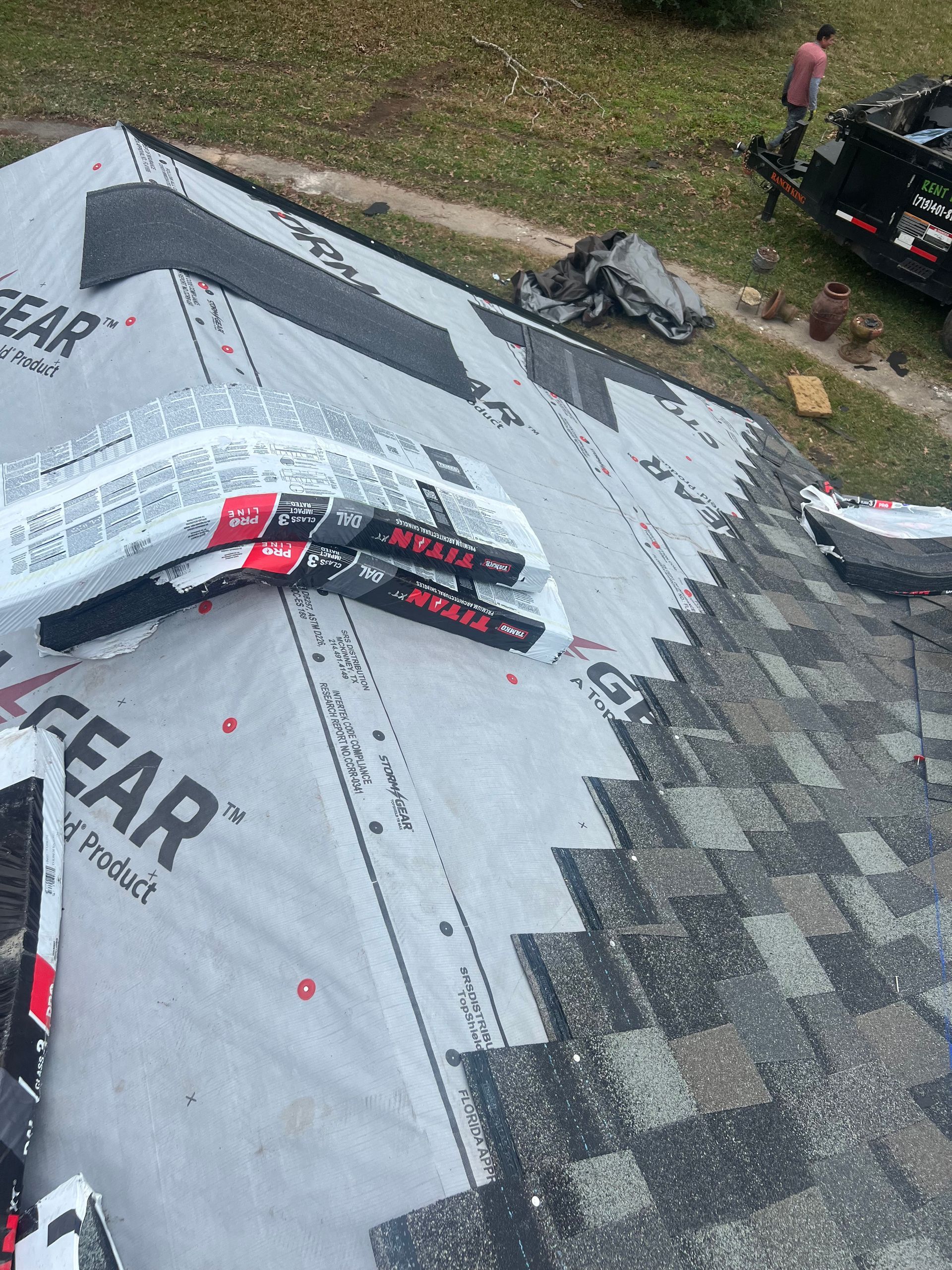 Roofing work in progress, asphalt shingles being laid. Rolls of shingles and a worker visible.