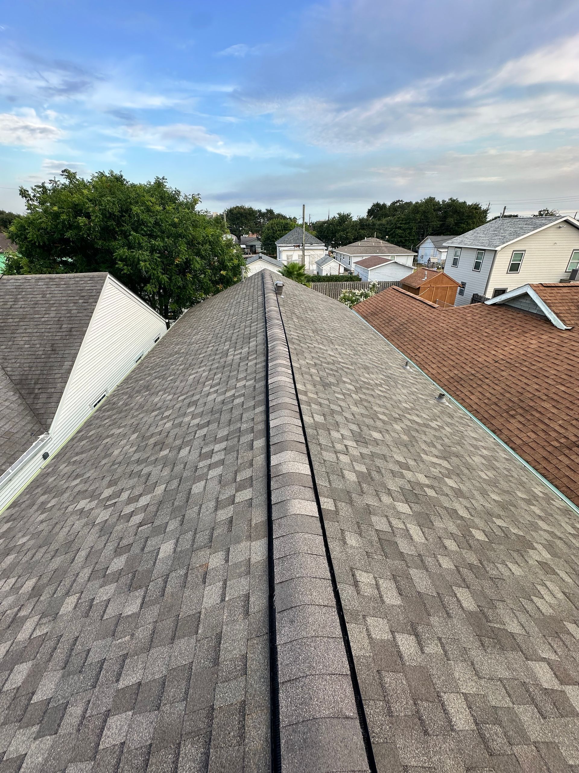 Gray asphalt shingle roof, view from peak, looking down a narrow street. Blue sky above houses.