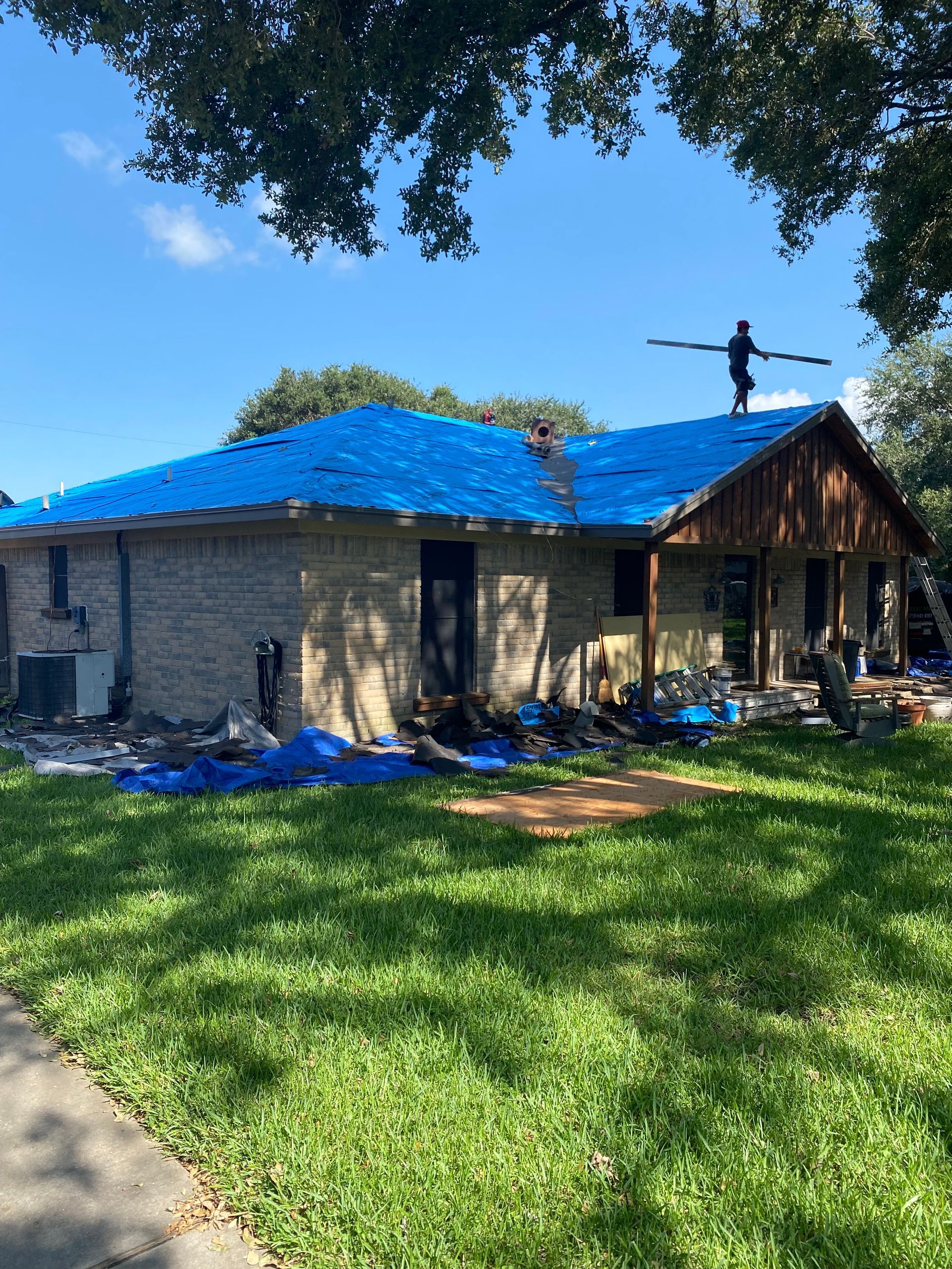 House with blue tarp on roof, workers. One worker on the roof holding wood pieces. Sunny day.