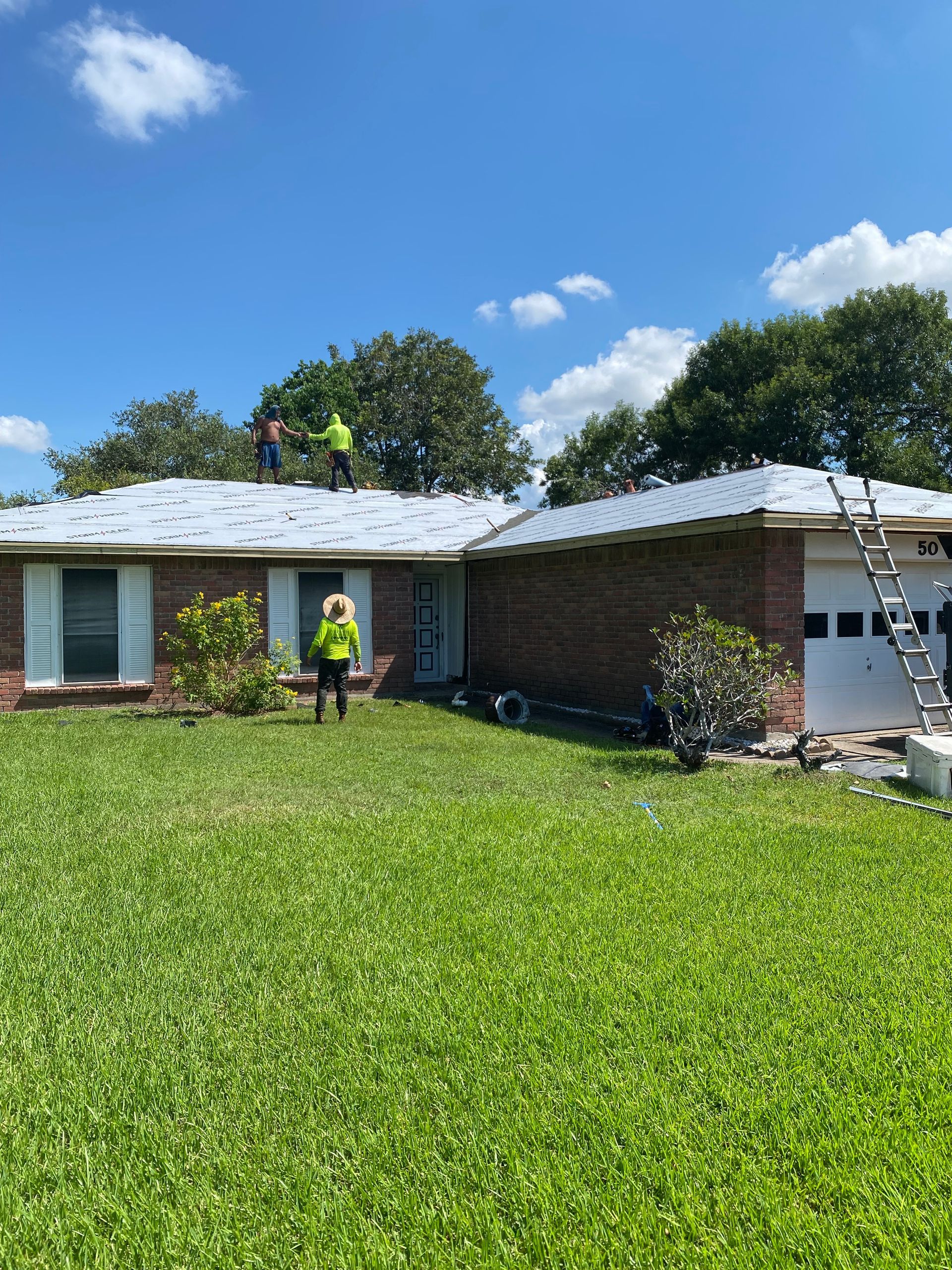 Two roofers on a house roof. A third person stands below. Green grass and blue sky.