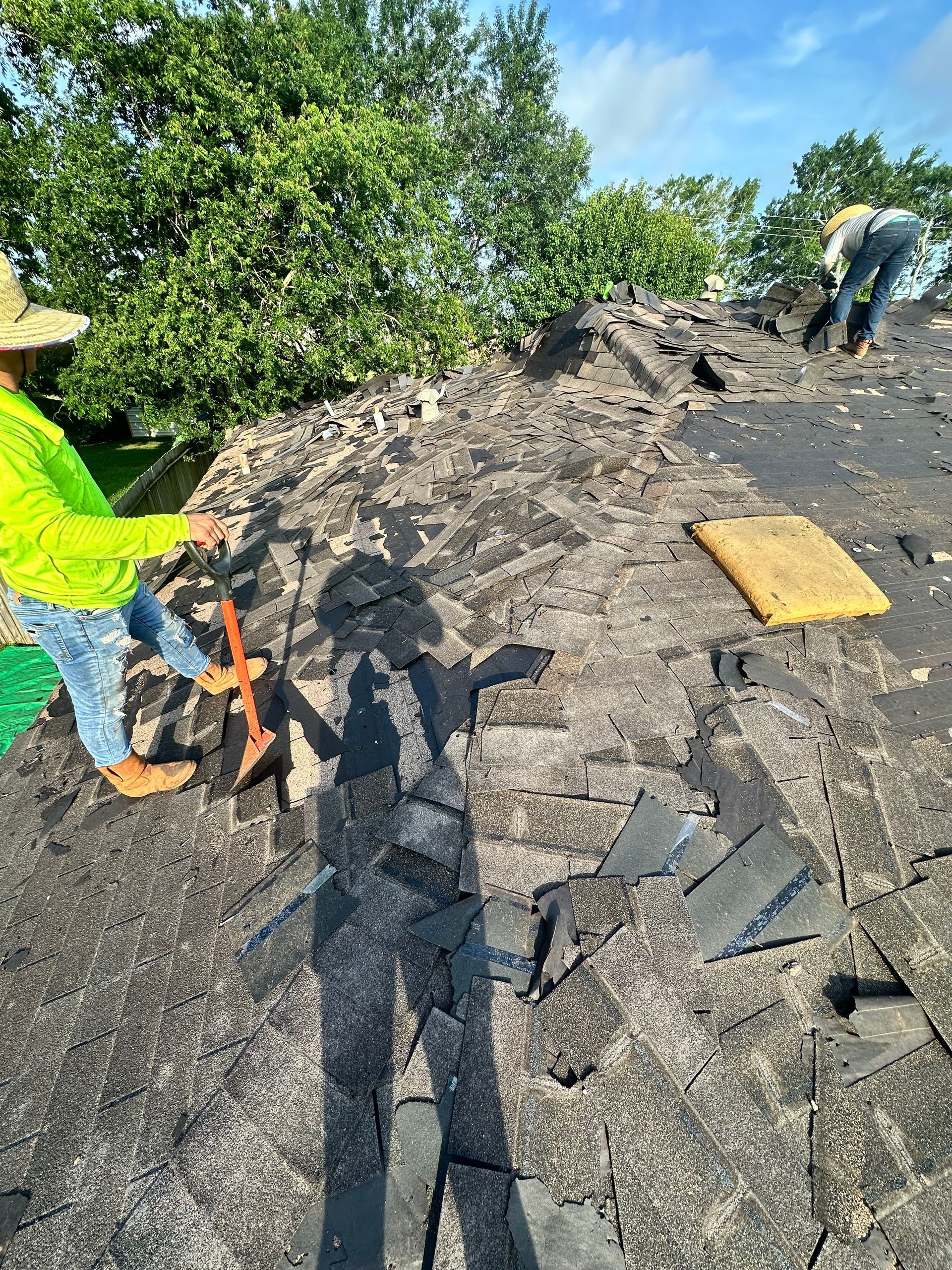 Workers removing shingles from a rooftop. One in neon green shirt, sunlight casting shadows.