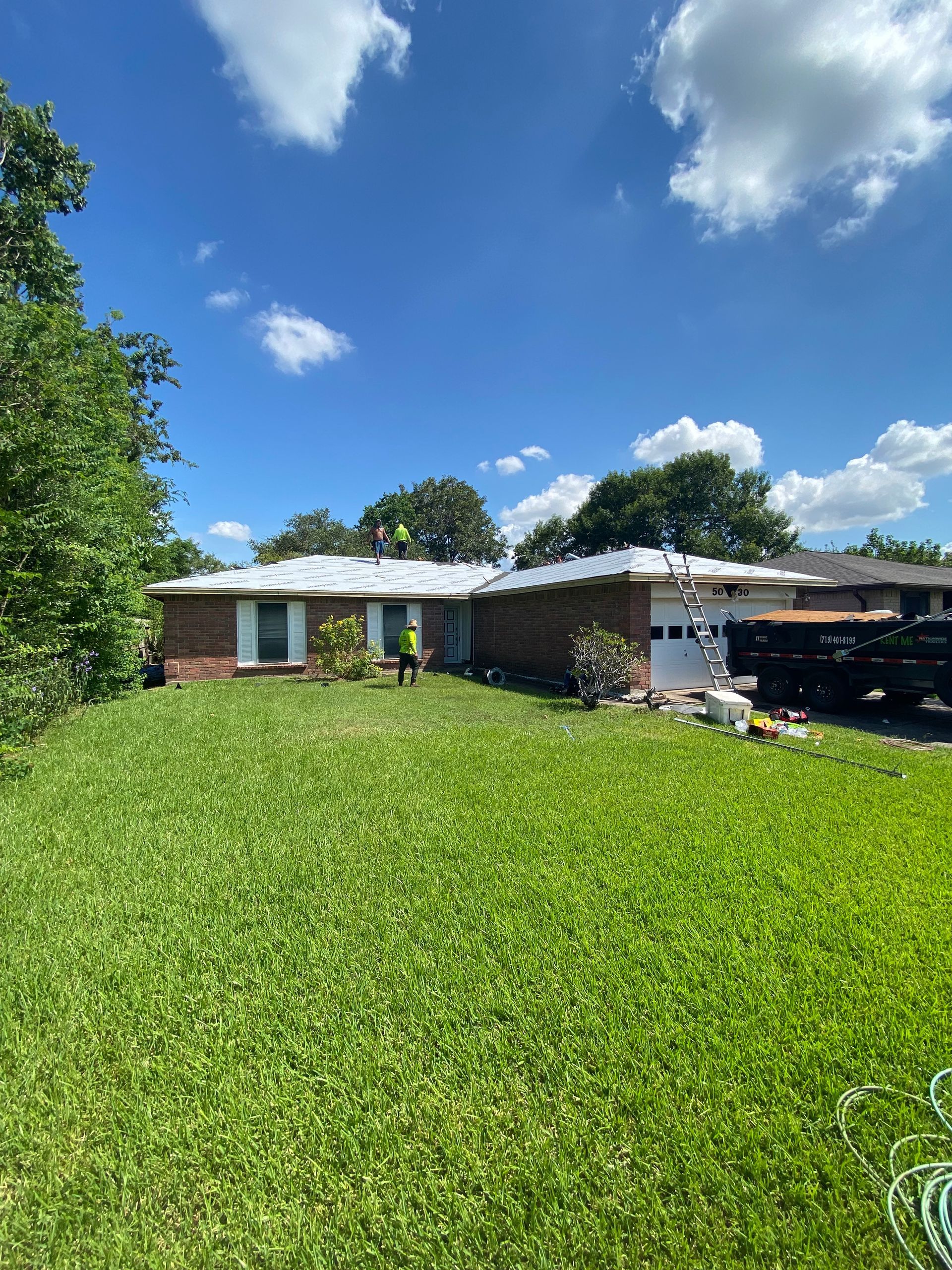 A brick house with roofers on the partially covered roof on a sunny day.
