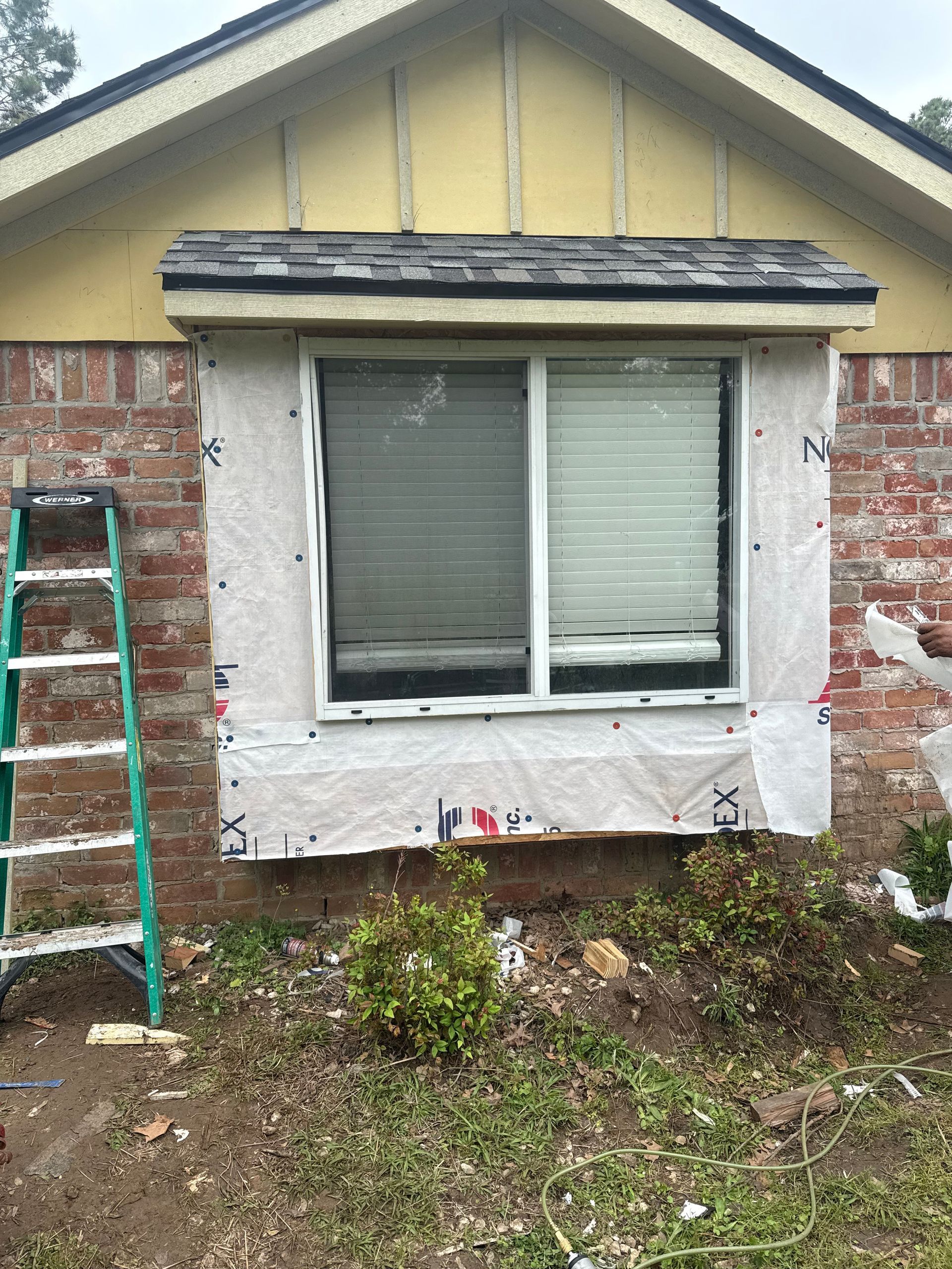 House exterior, window replacement in progress. Brick wall, white window frame, protective wrap, small plants, and ladder.