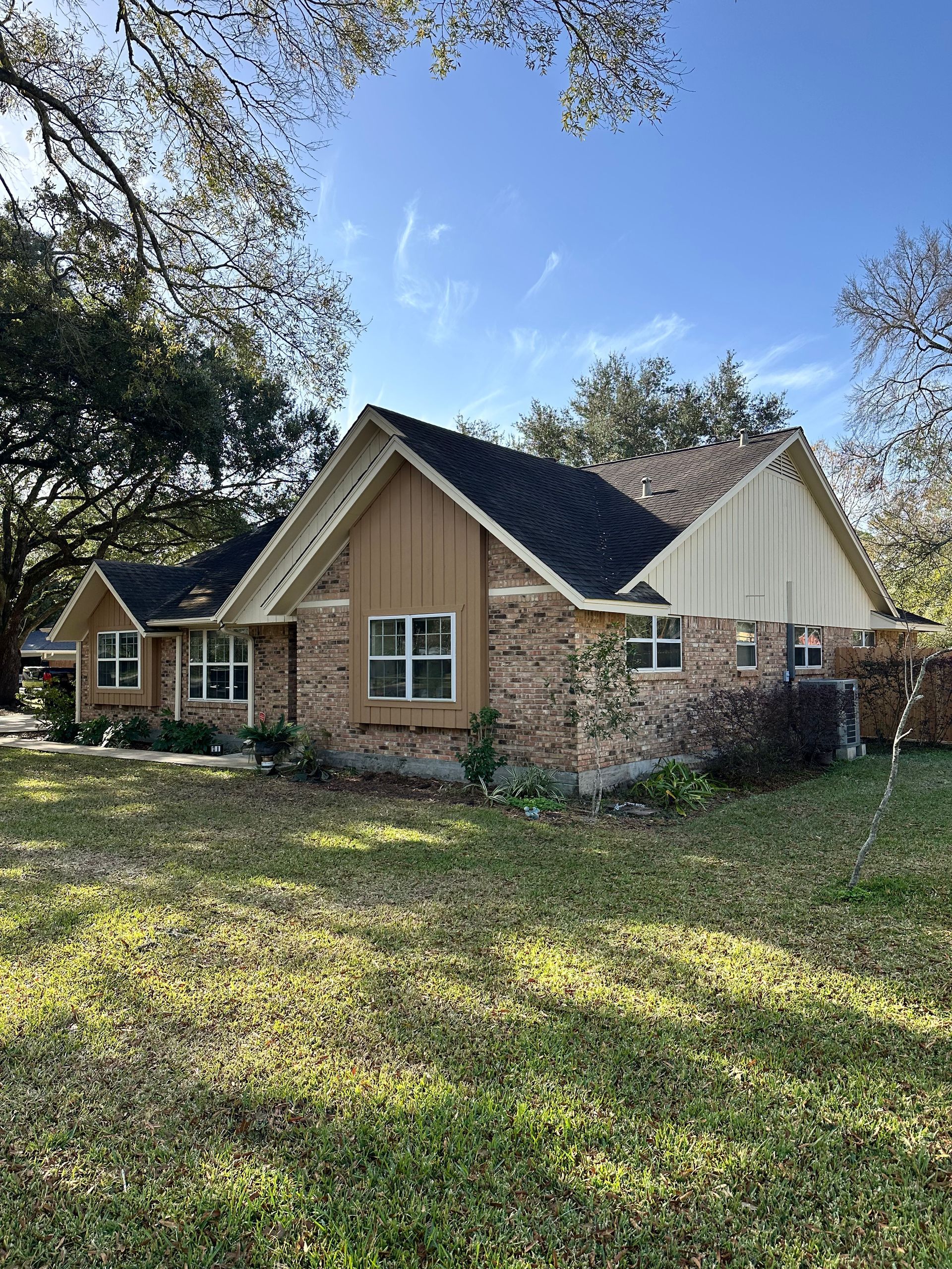 Brick house with a brown roof and tan siding, framed by trees under a blue sky.