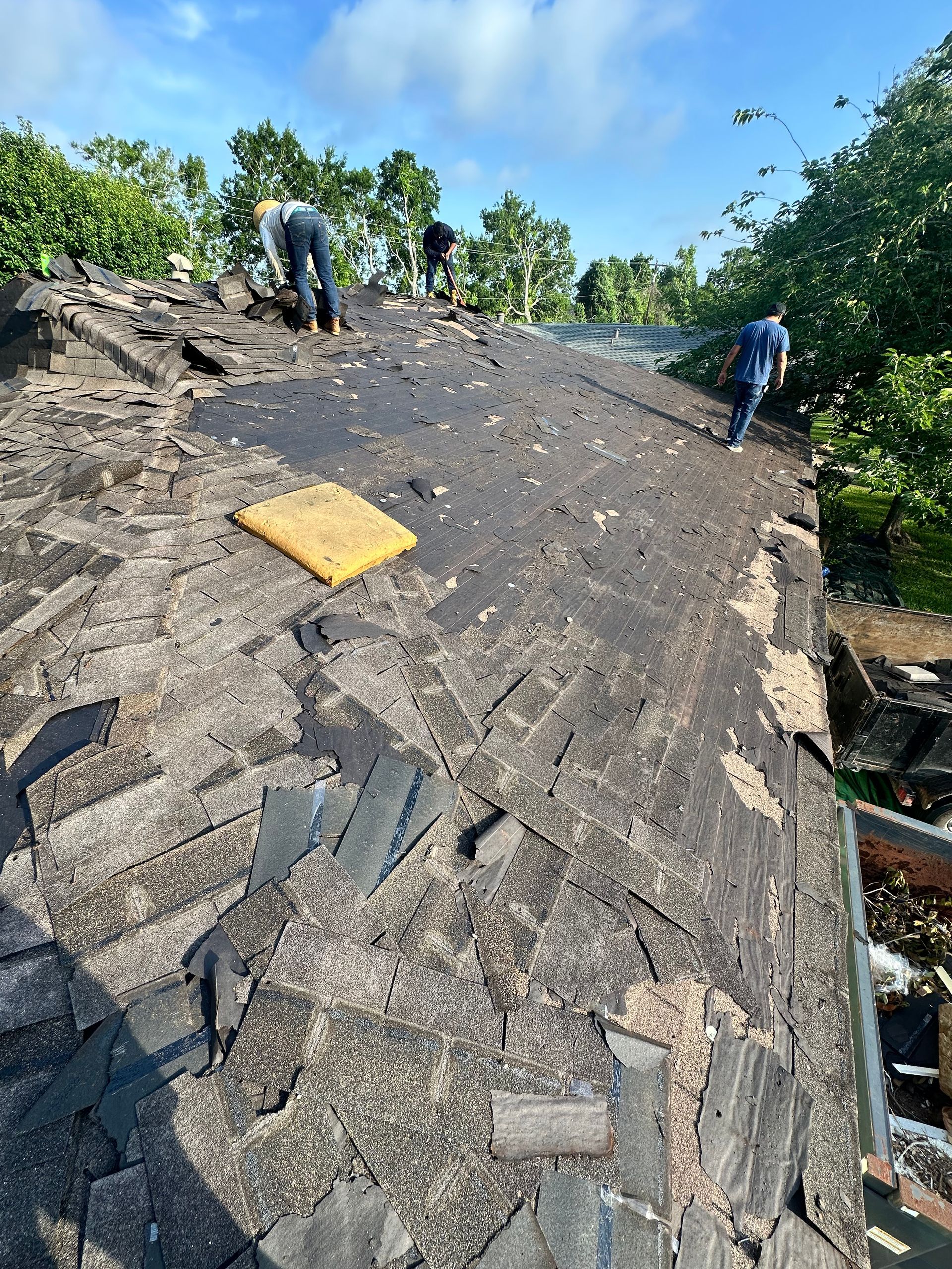Workers on a damaged roof, tearing off old shingles. Blue sky and green trees in background.