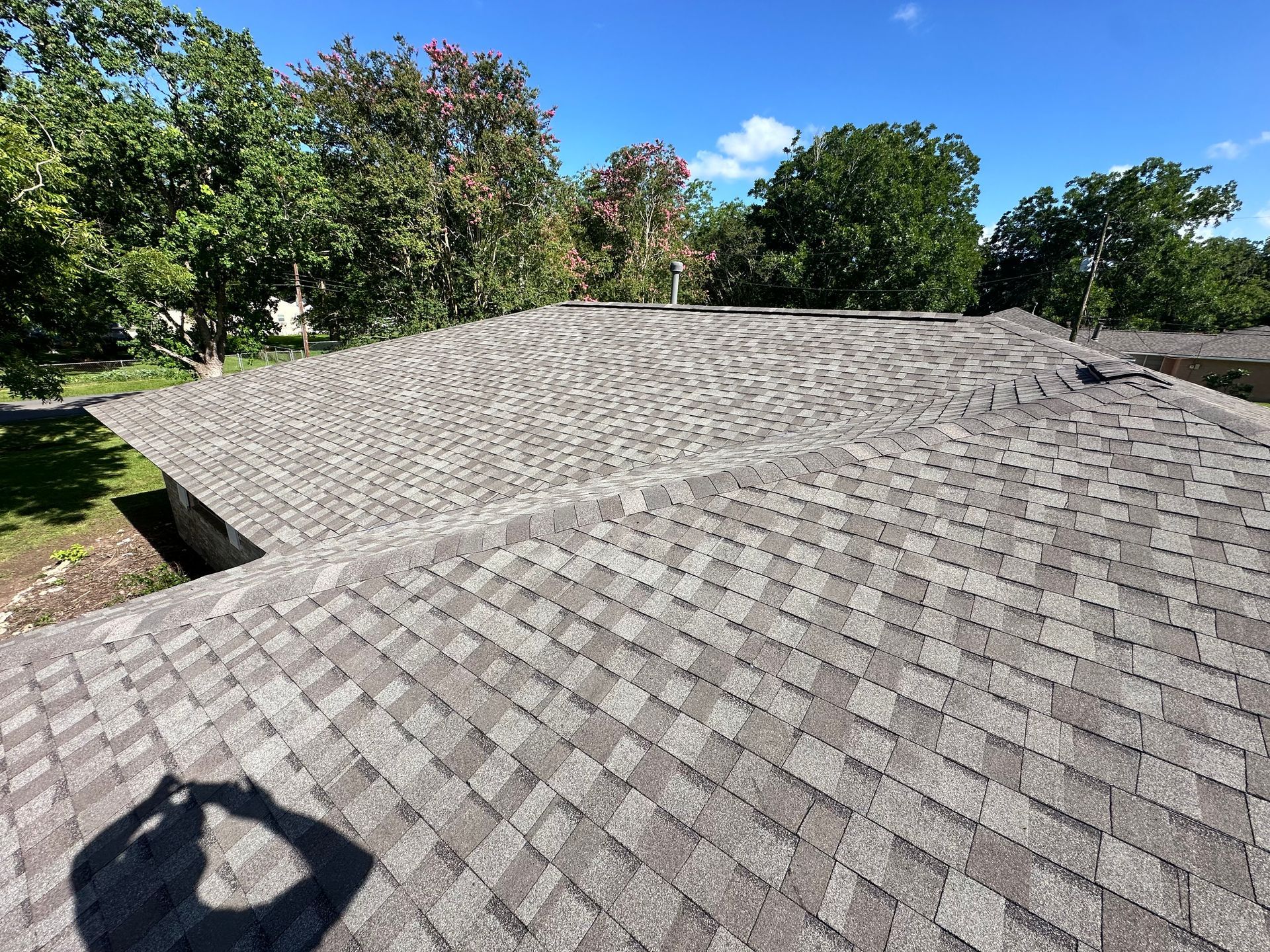 Gray shingle roof on a house with trees in the background under a blue sky.