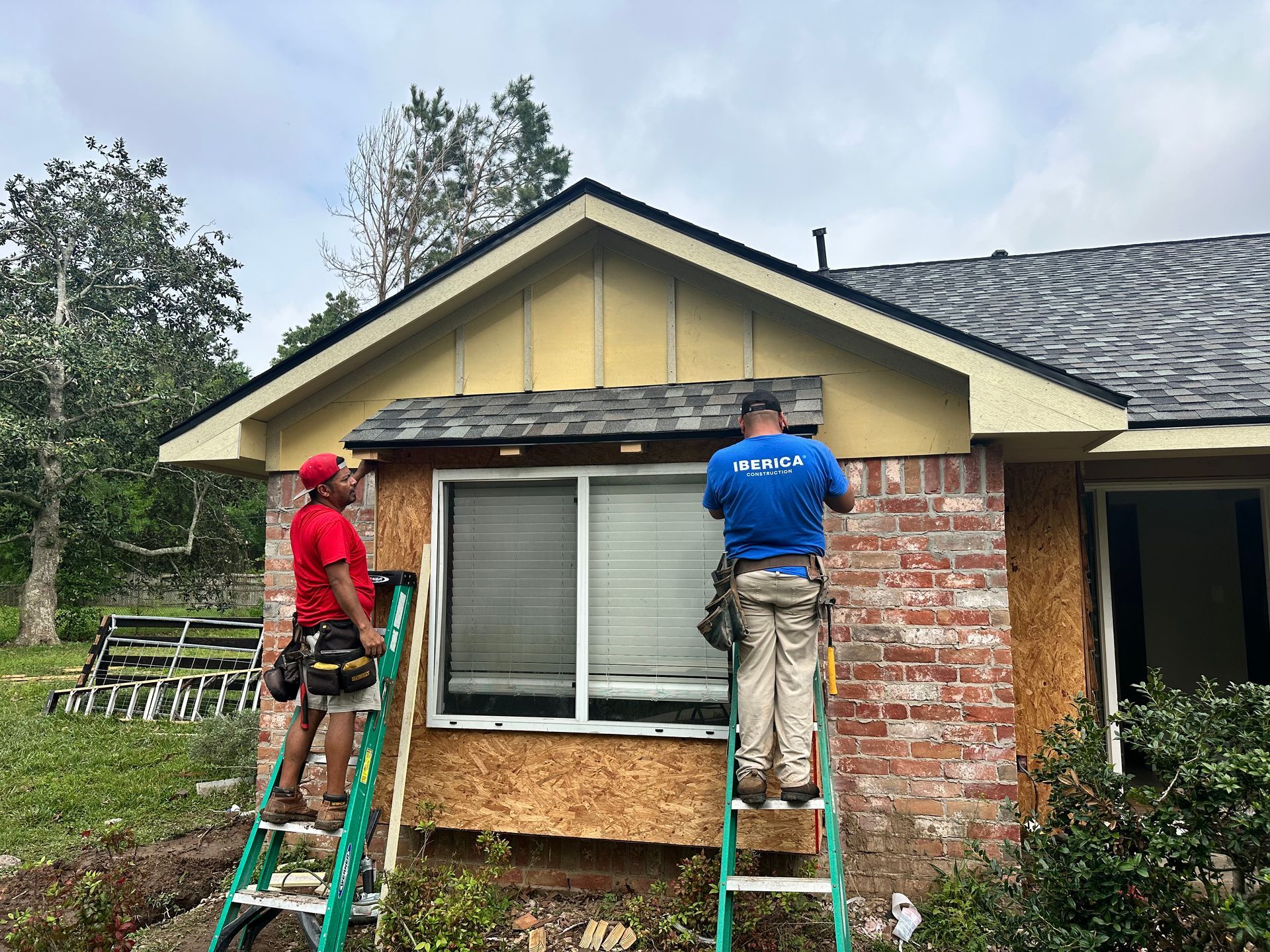 Two workers on ladders repair the exterior of a brick house. One wears red, the other blue, working on the siding near a window.
