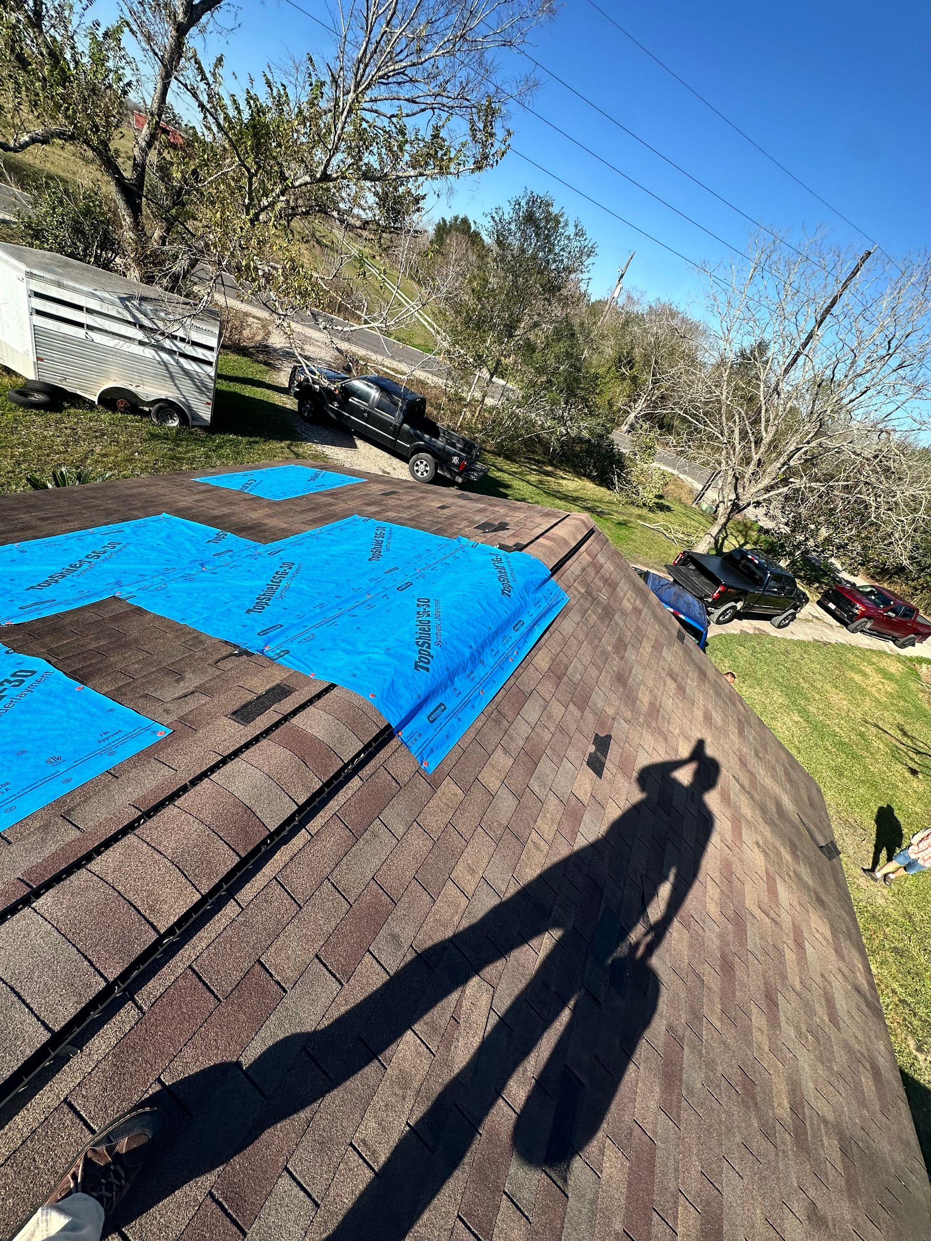 Rooftop with blue tarps. Shadow of person holding a bag. Brown shingles, trees, blue sky.