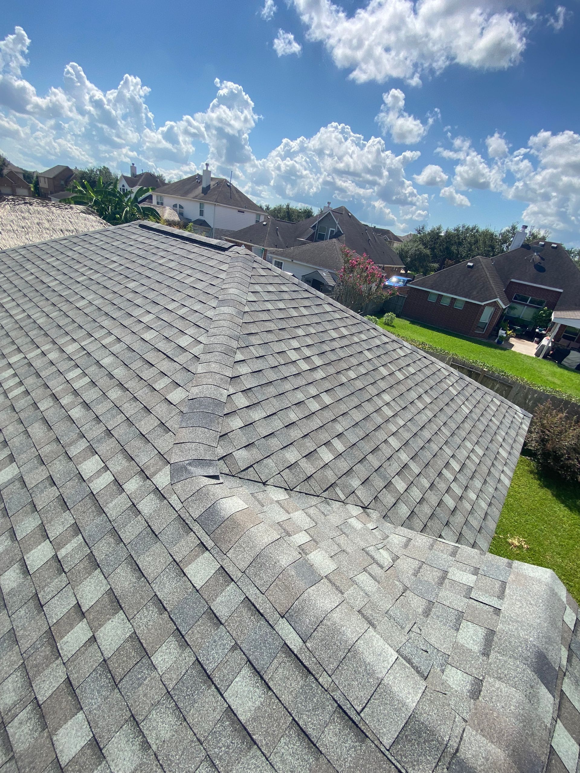 Overhead view of a gray shingle roof under a blue sky with fluffy white clouds; grass and houses in the background.