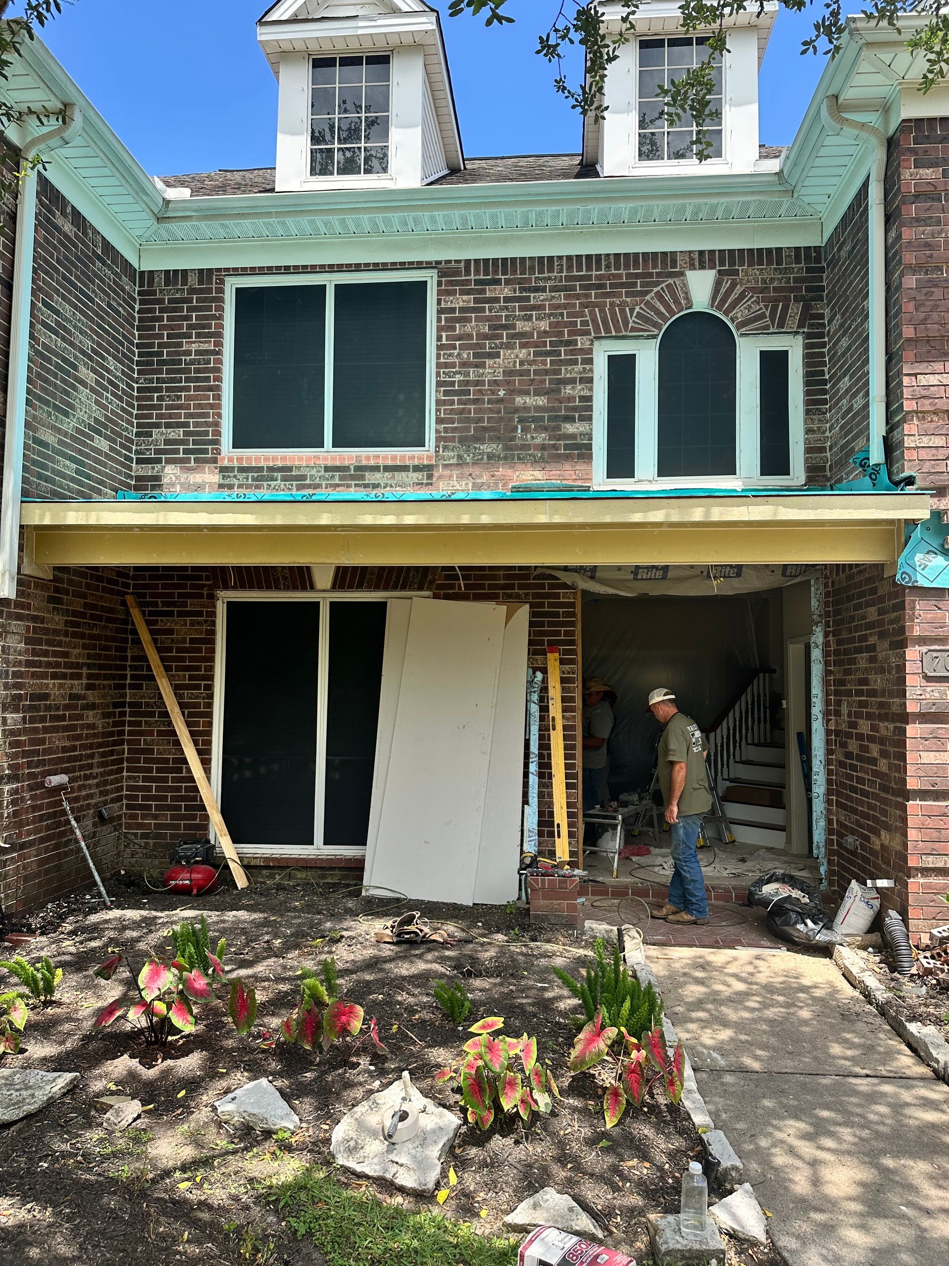 A house undergoing renovation, with exposed framing, workers, and debris. Brick exterior, green trim, and plants in foreground.
