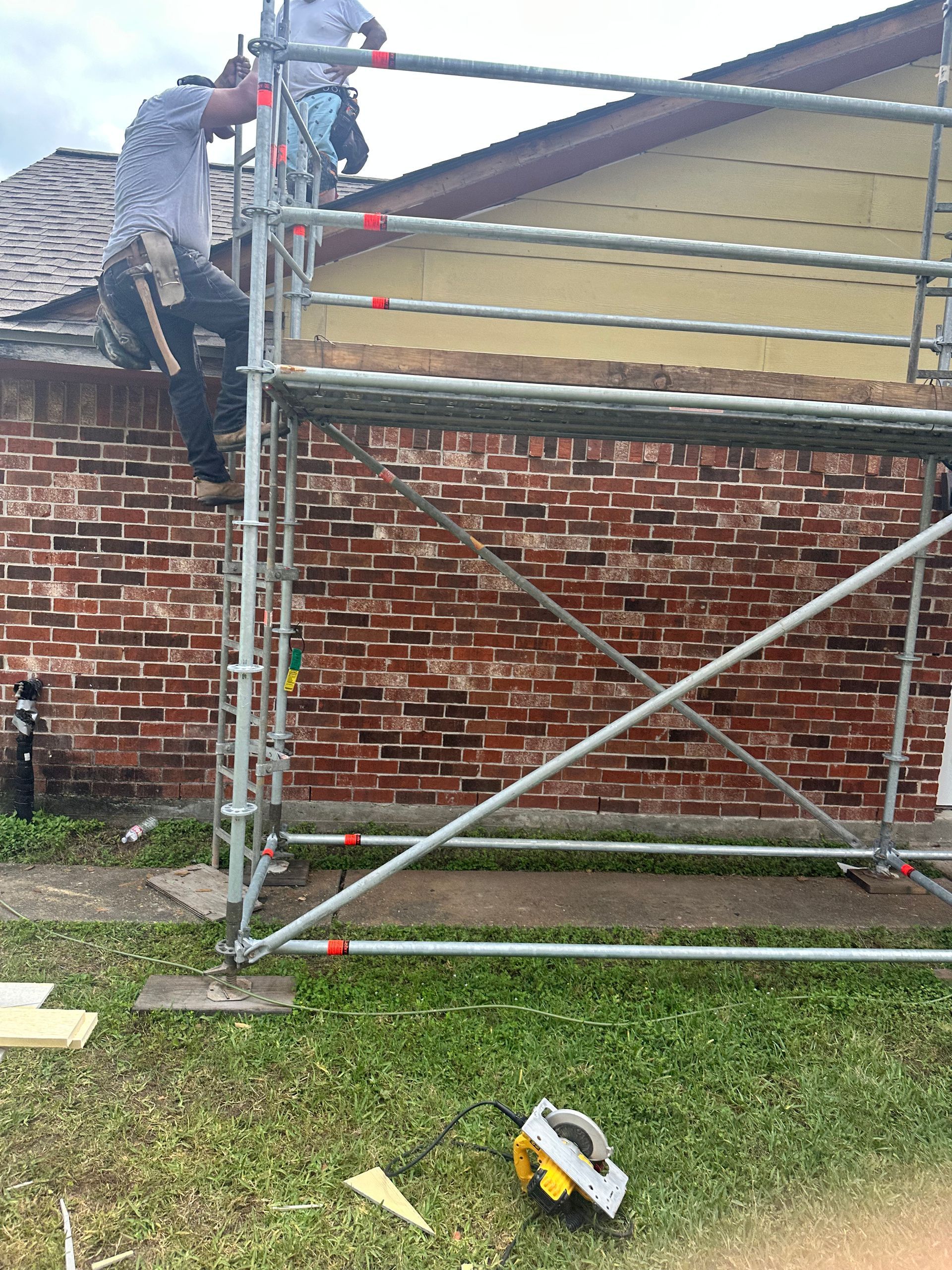 Two construction workers on scaffolding near a brick wall and yellow siding, one working on the roof.